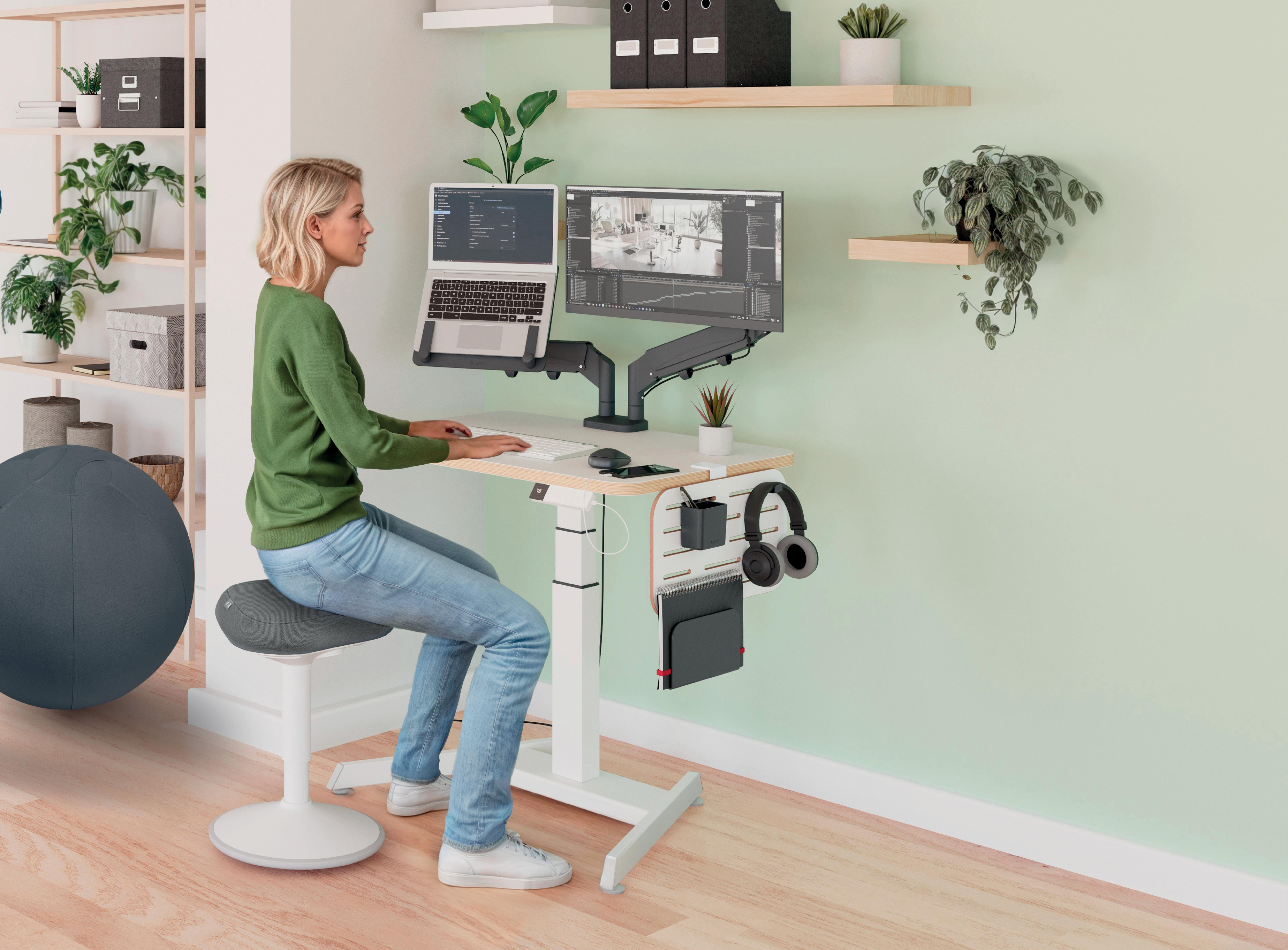 Frau an einem Schreibtisch mit Laptop und Monitor, auf einem Hocker sitzend. Grüne Wand und Regale im Hintergrund.