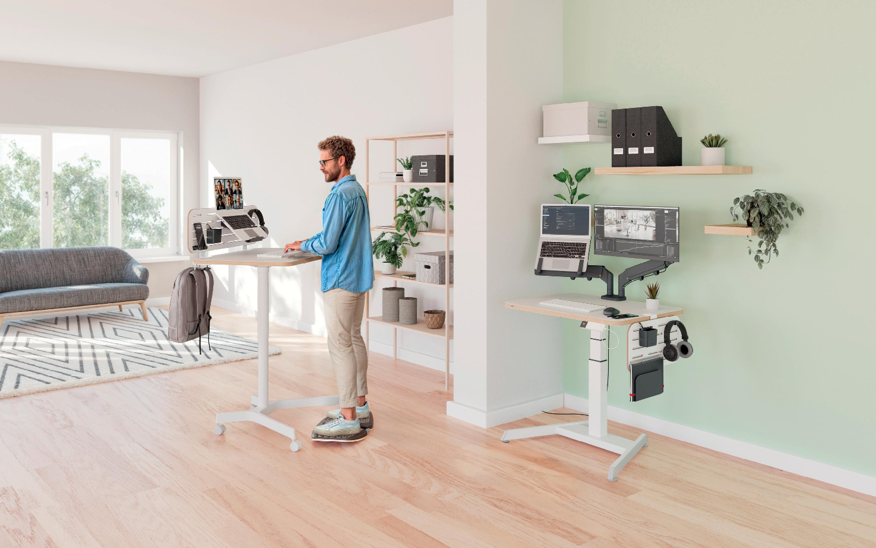 Ein Mann steht an einem Stehpult in einem Homeoffice mit zwei Monitoren. Es gibt eine Couch, Regale und Pflanzen.