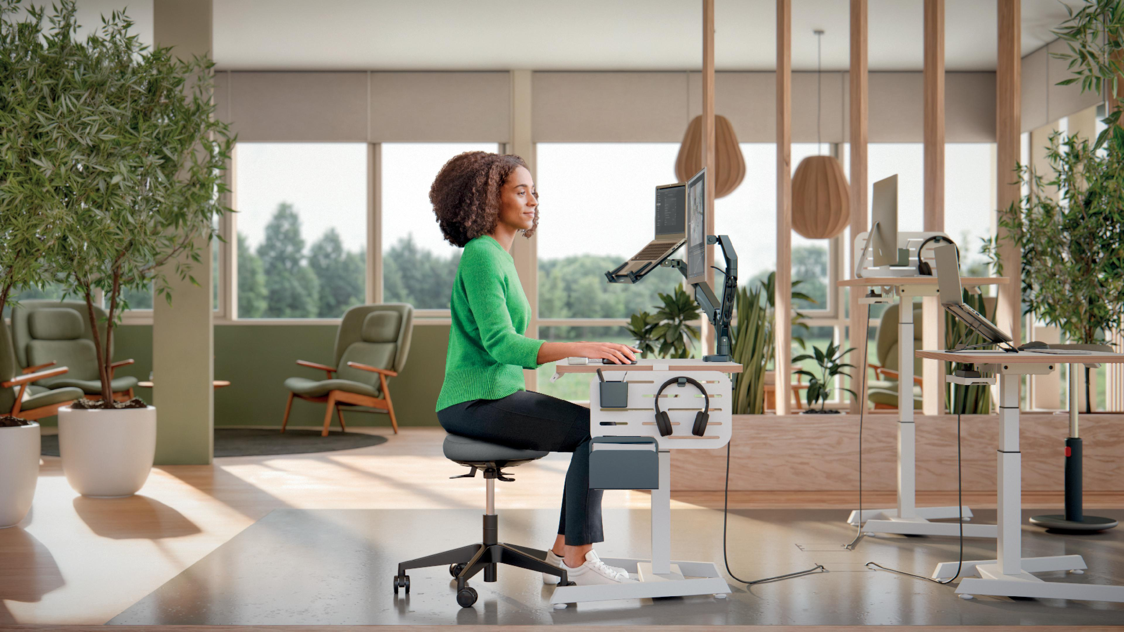 Eine Frau in Grün sitzt an einem modernen Schreibtisch mit Laptop und Monitor. Pflanzen, Fenster und Stühle sind im Hintergrund.
