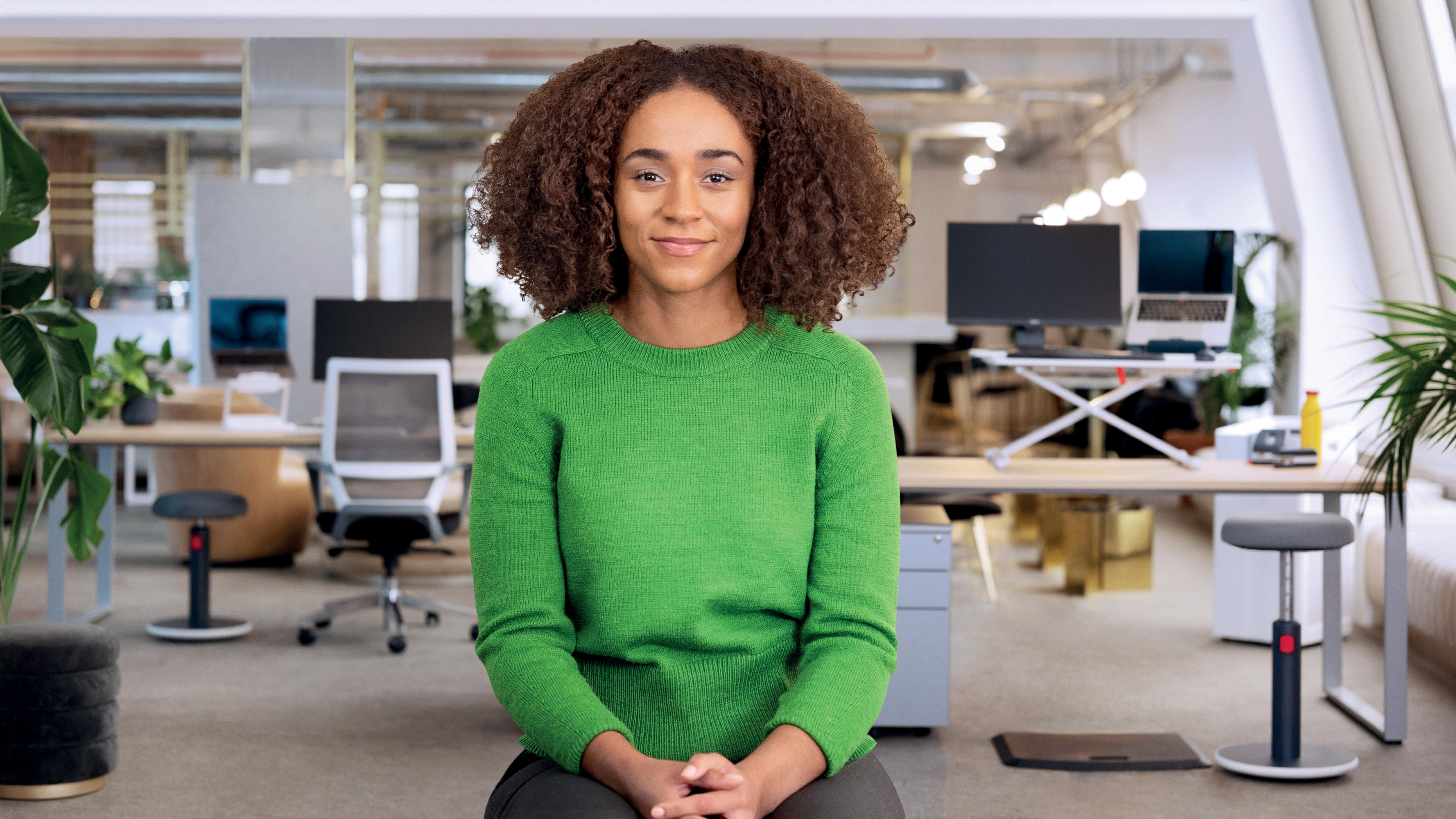 Eine Frau mit lockigem Haar lächelt in einem Büro. Sie trägt einen grünen Pullover. Im Hintergrund sind Schreibtische, Pflanzen und Computer.