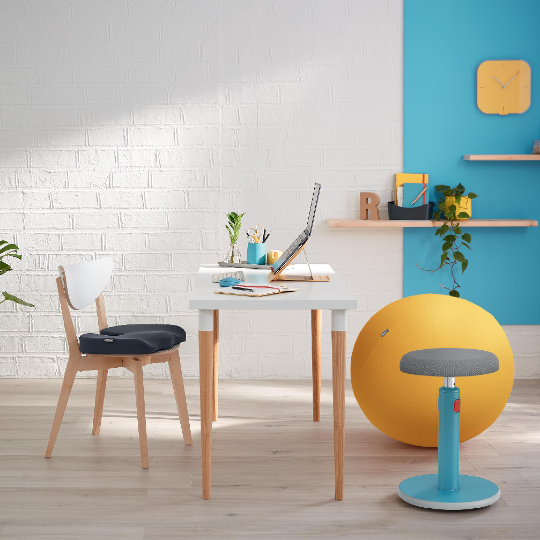 Schreibtisch mit Laptop, Stuhl und gelbem Gymnastikball. Blaue und weiße Wände, Holzboden.