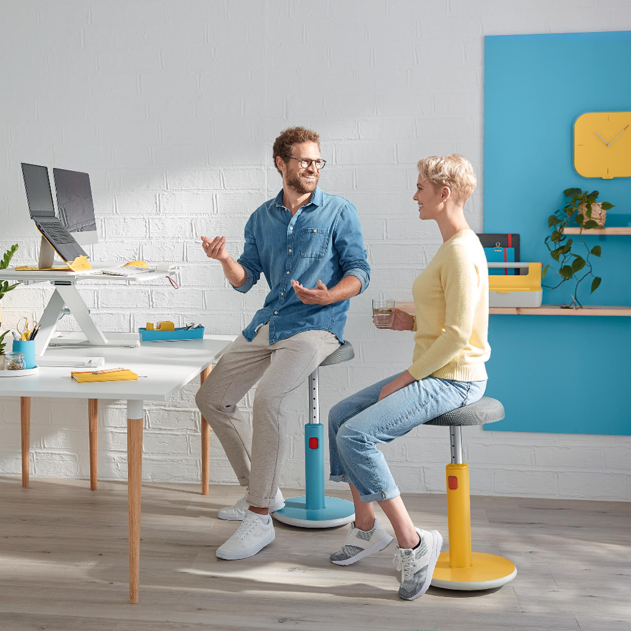Zwei Personen sitzen auf Hockern und diskutieren. Ein Schreibtisch mit Laptops und Schreibwaren. Weißer Ziegelhintergrund.