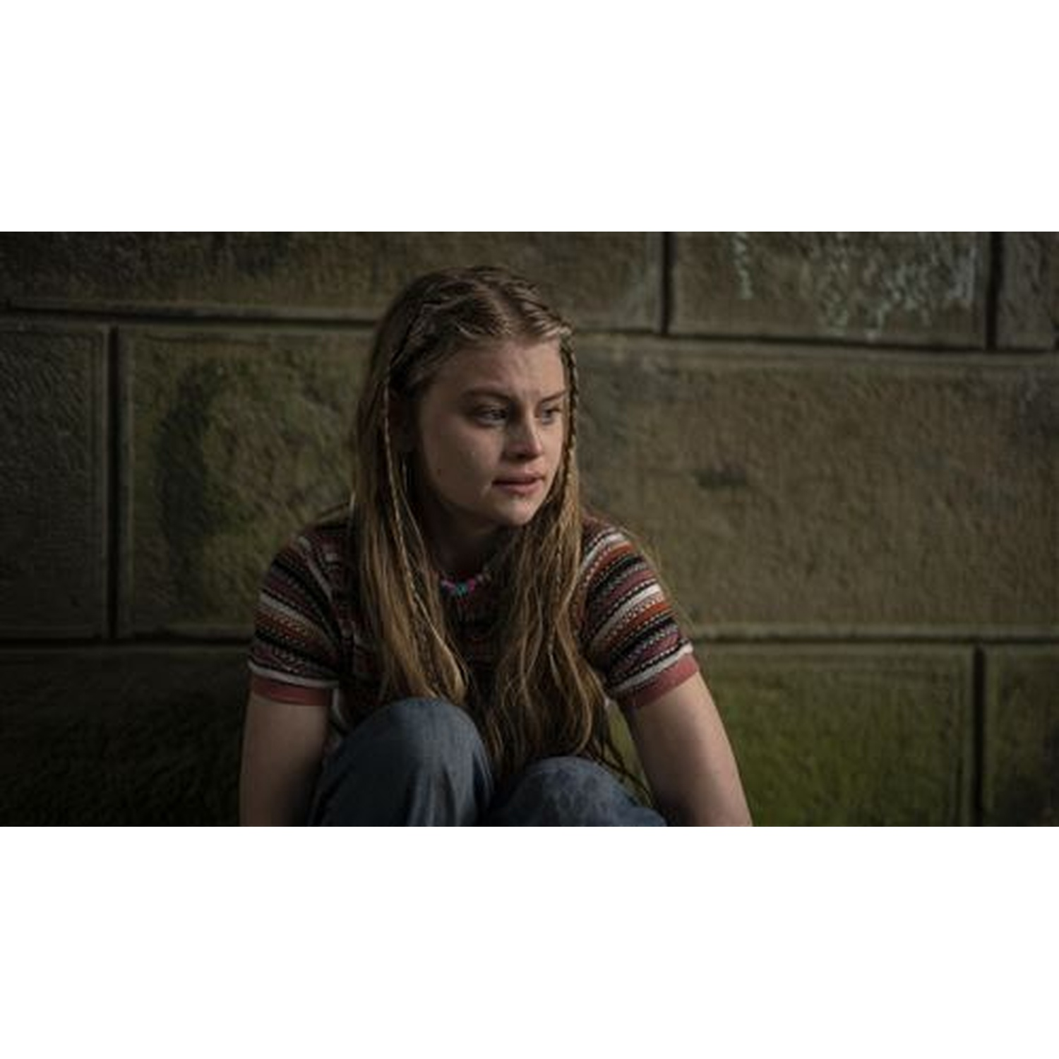Een jonge vrouw met lang haar en een shirt met patroon zit tegen een stenen muur. Ze lijkt bedachtzaam, met een ketting.