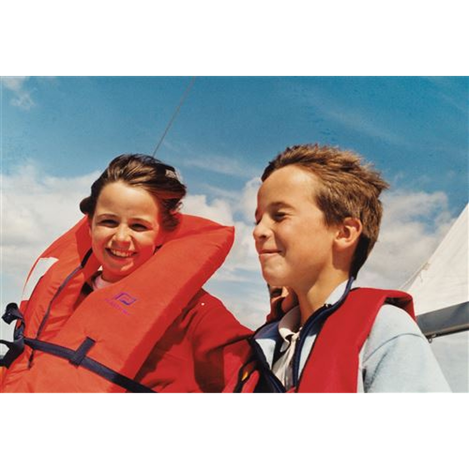 Deux enfants portant des gilets de sauvetage rouges sourient sur un bateau, sous un ciel bleu nuageux. L'un a les cheveux foncés, l'autre plus clairs.
