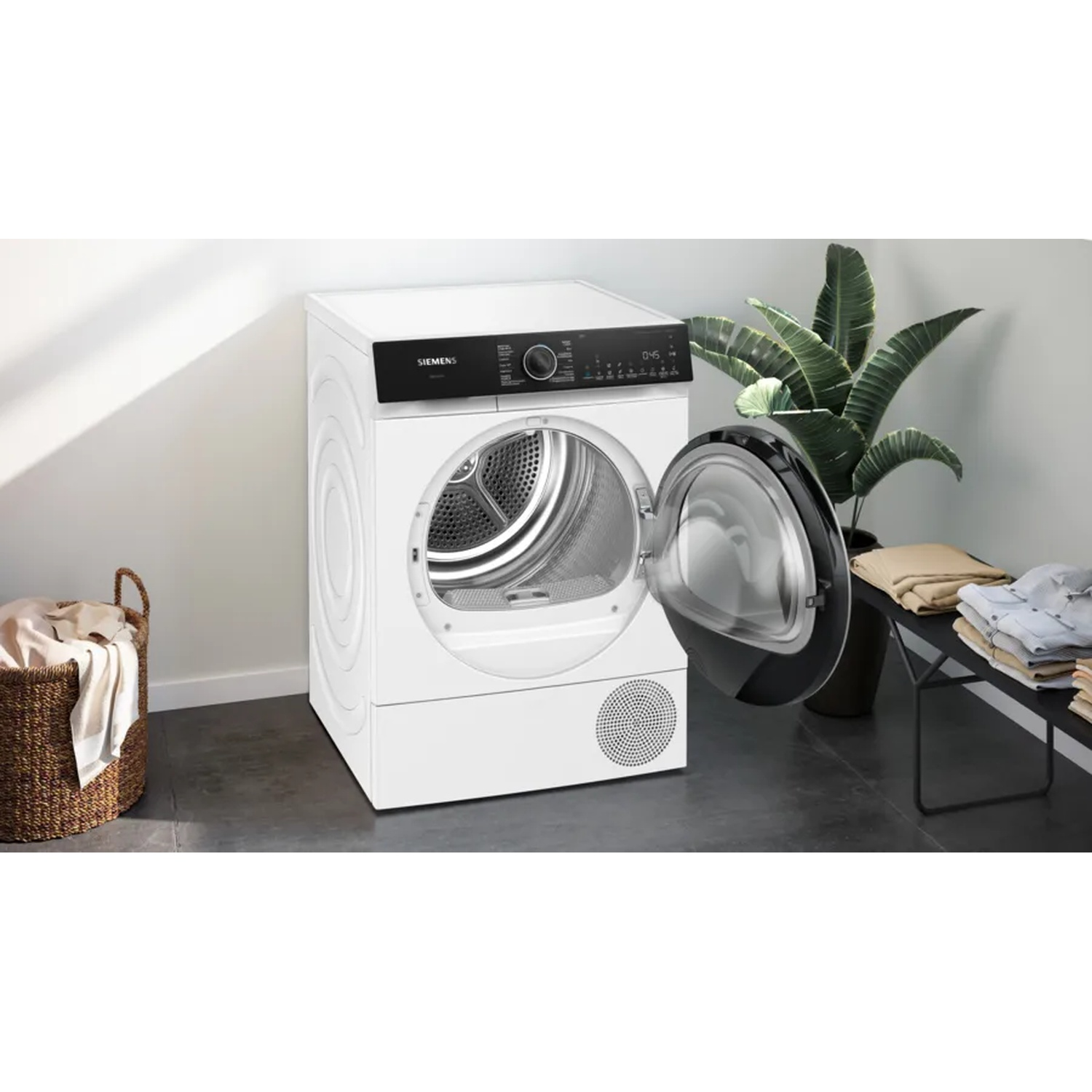 A white Siemens dryer with the door open. A basket of clothes is on the left, and plants and folded clothes are on the right.