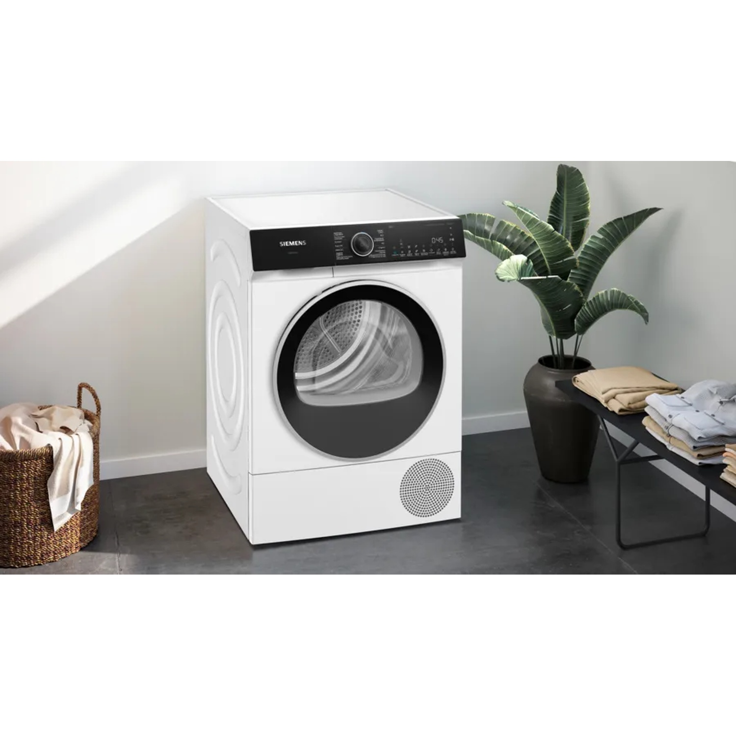A white Siemens dryer with a black door, a basket of clothes, a potted plant, and folded clothes on a black table.