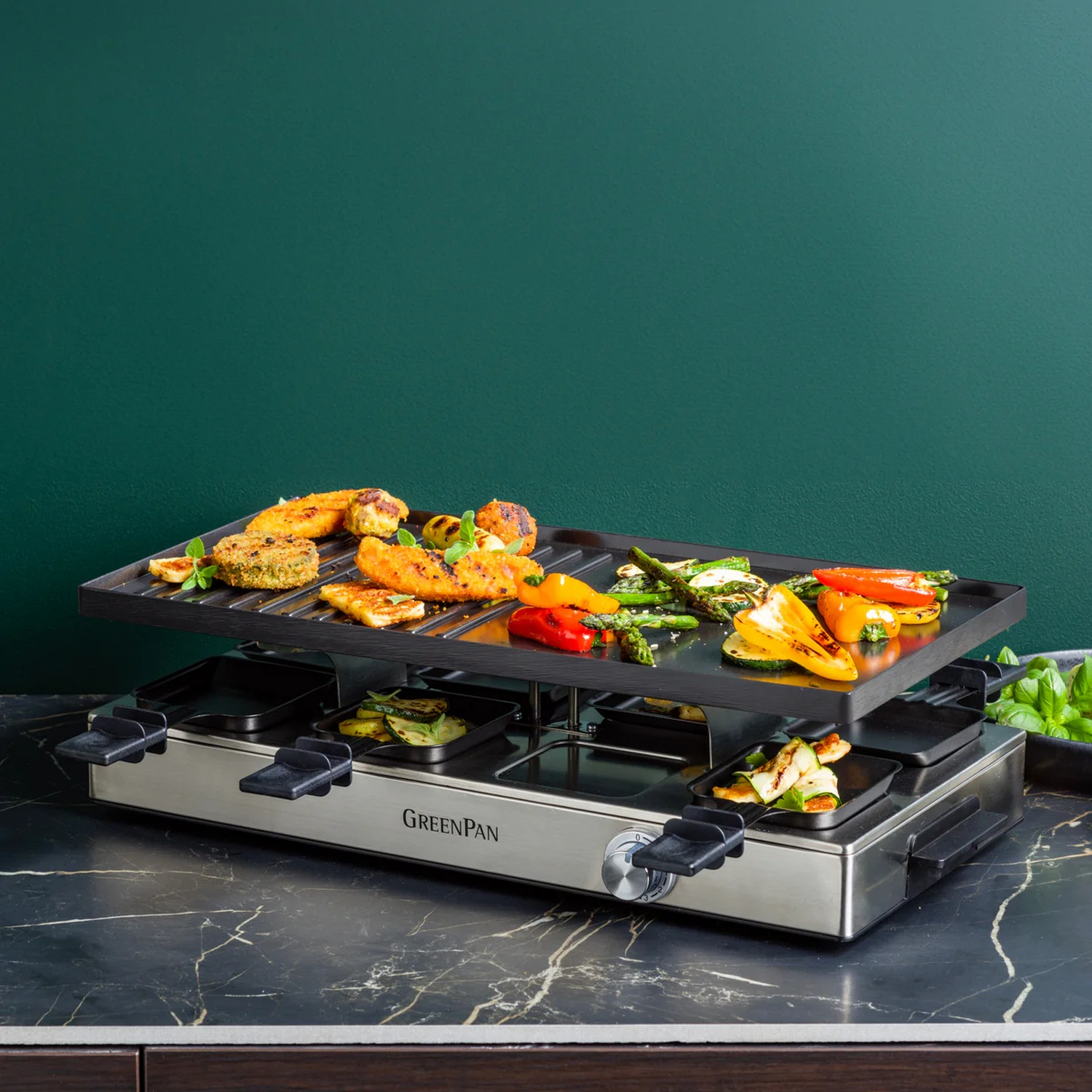 Raclette grill met gegrilde groenten en gefrituurd eten erop. Groene muur op de achtergrond. Zwart marmeren aanrecht.