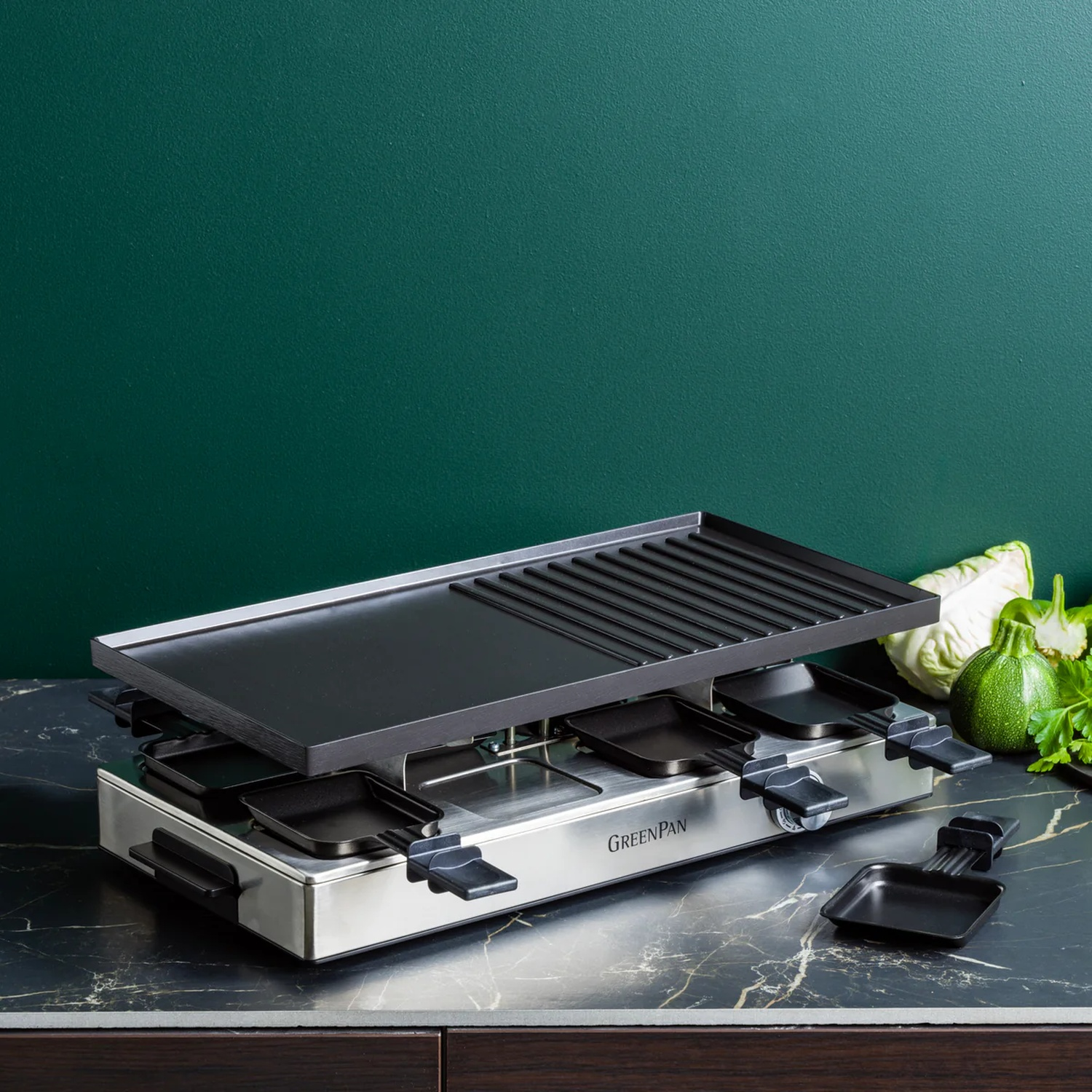 Raclette grill met zwarte grillplaat en pannen, op een marmeren aanrecht. Groene groenten op de achtergrond.