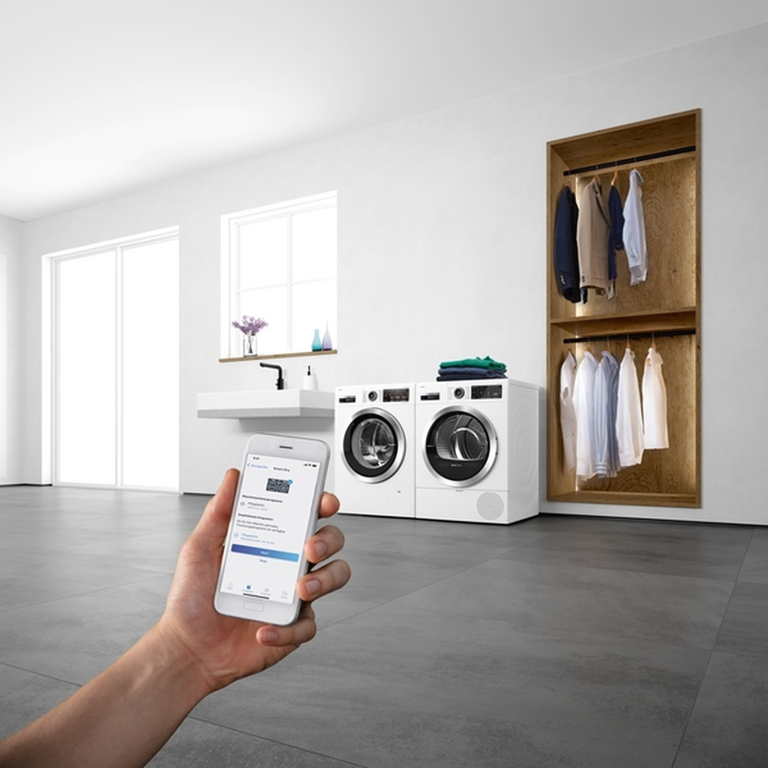 Une personne tenant un smartphone est dans une buanderie avec une laveuse, une sécheuse et un placard. La pièce est moderne.