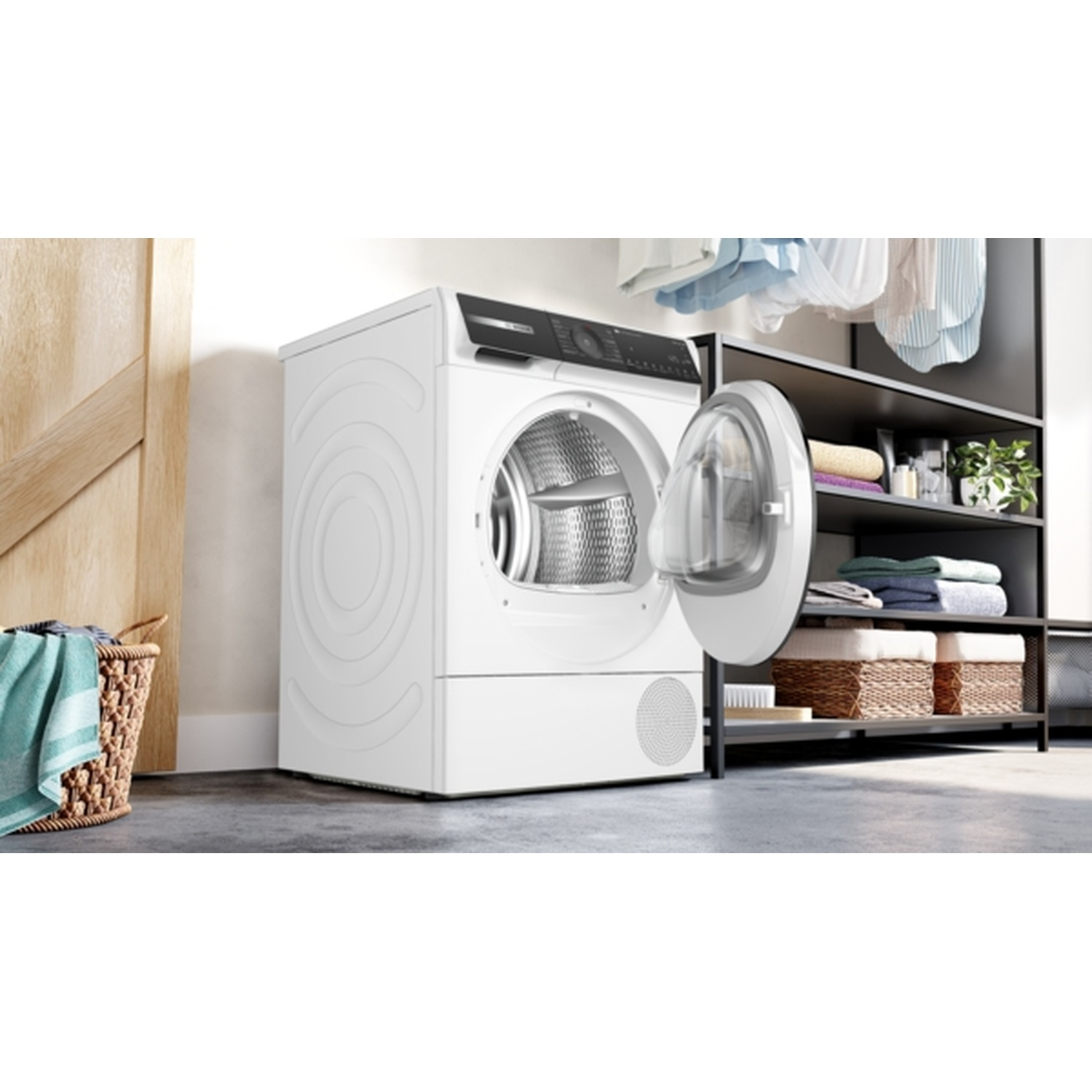 Un sèche-linge blanc avec la porte ouverte, une porte en bois, un panier à linge et des étagères avec des serviettes et des paniers sont visibles.