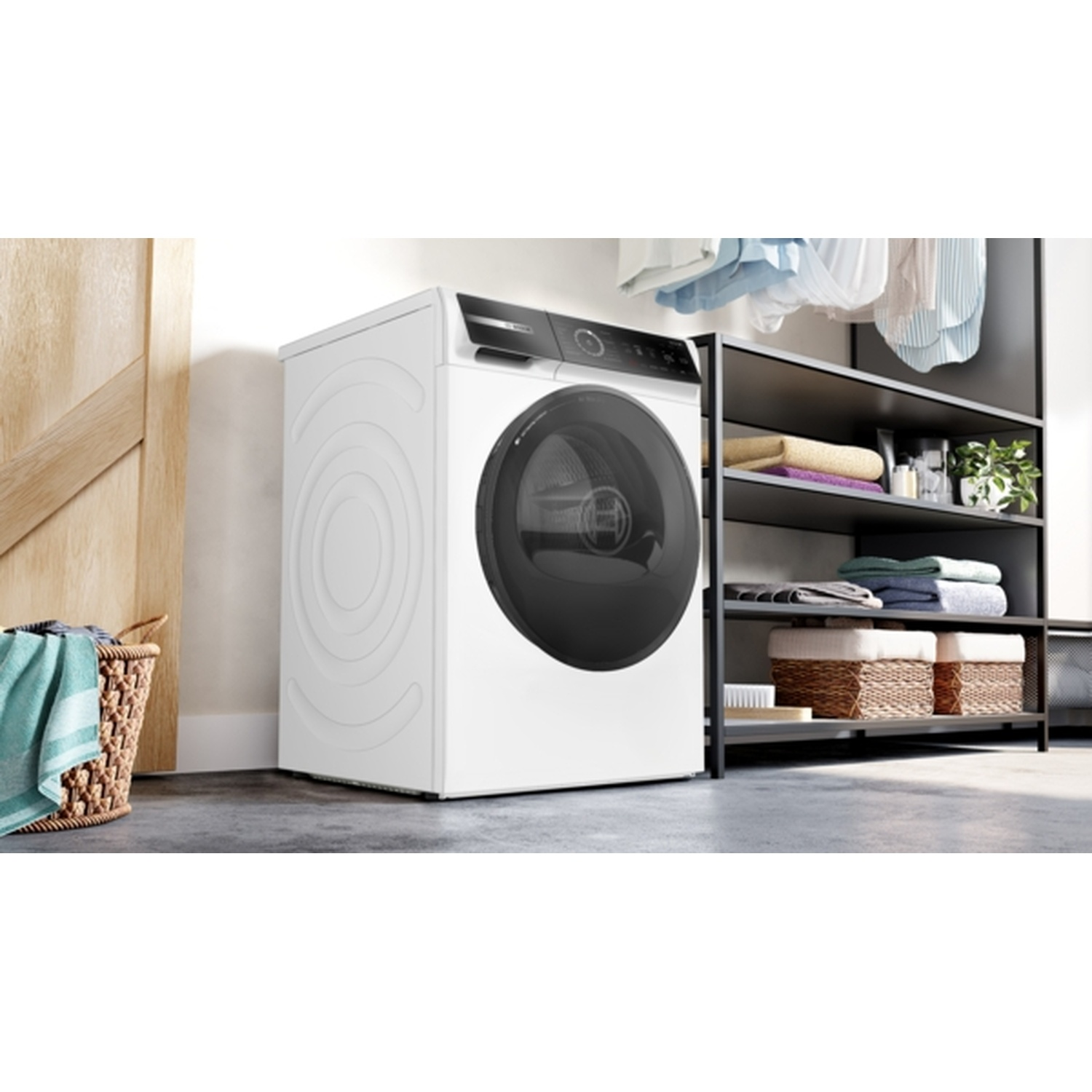 A white washing machine with a black door stands next to a shelf with folded towels and laundry baskets. Clothes hang above.