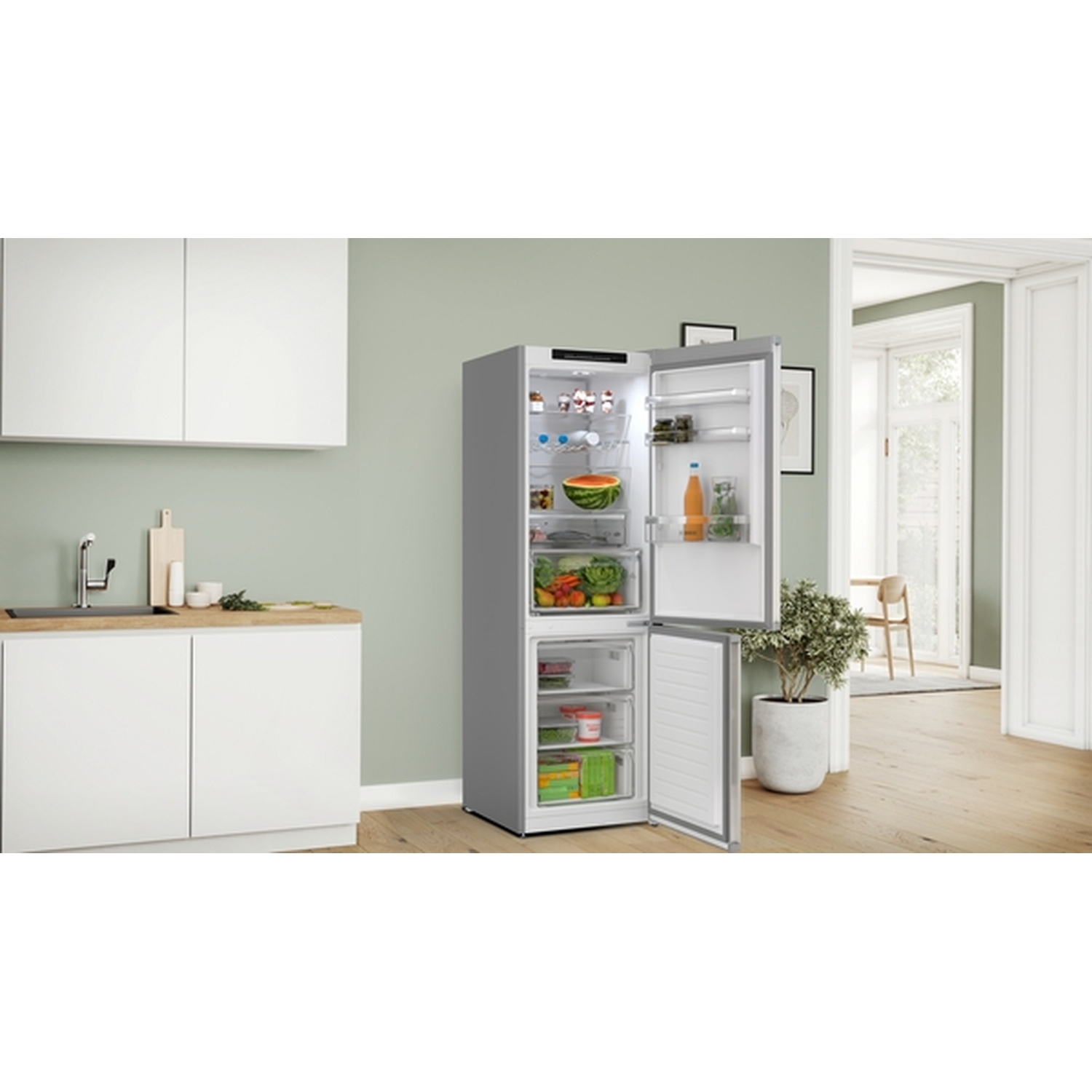 Open koelkast in een keuken vol eten. Witte kasten en een plant links. Tafel en stoelen op de achtergrond.