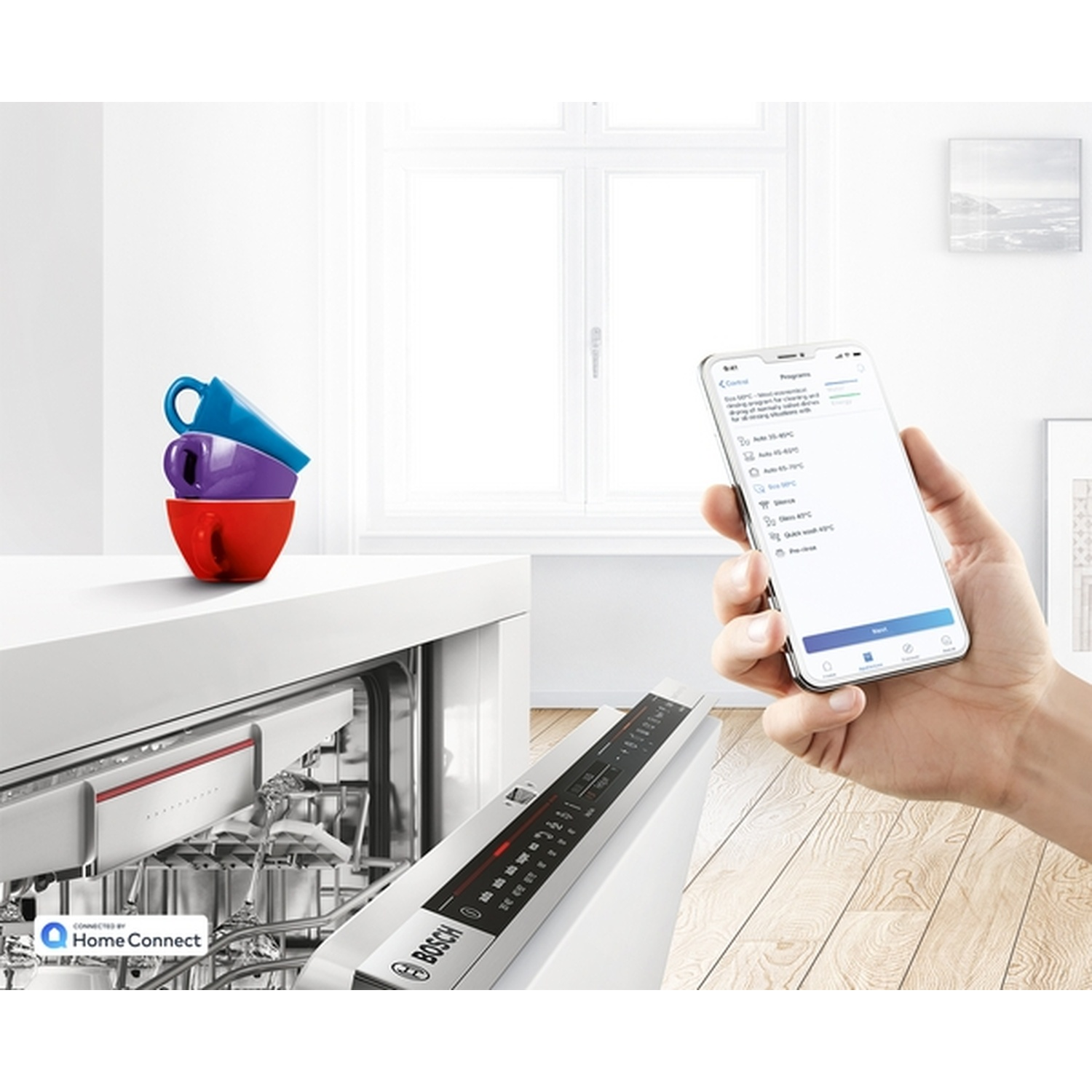 Une main tenant un smartphone affichant les paramètres du lave-vaisselle. Tasses rouges, violettes et bleues empilées. Un lave-vaisselle Bosch en arrière-plan.