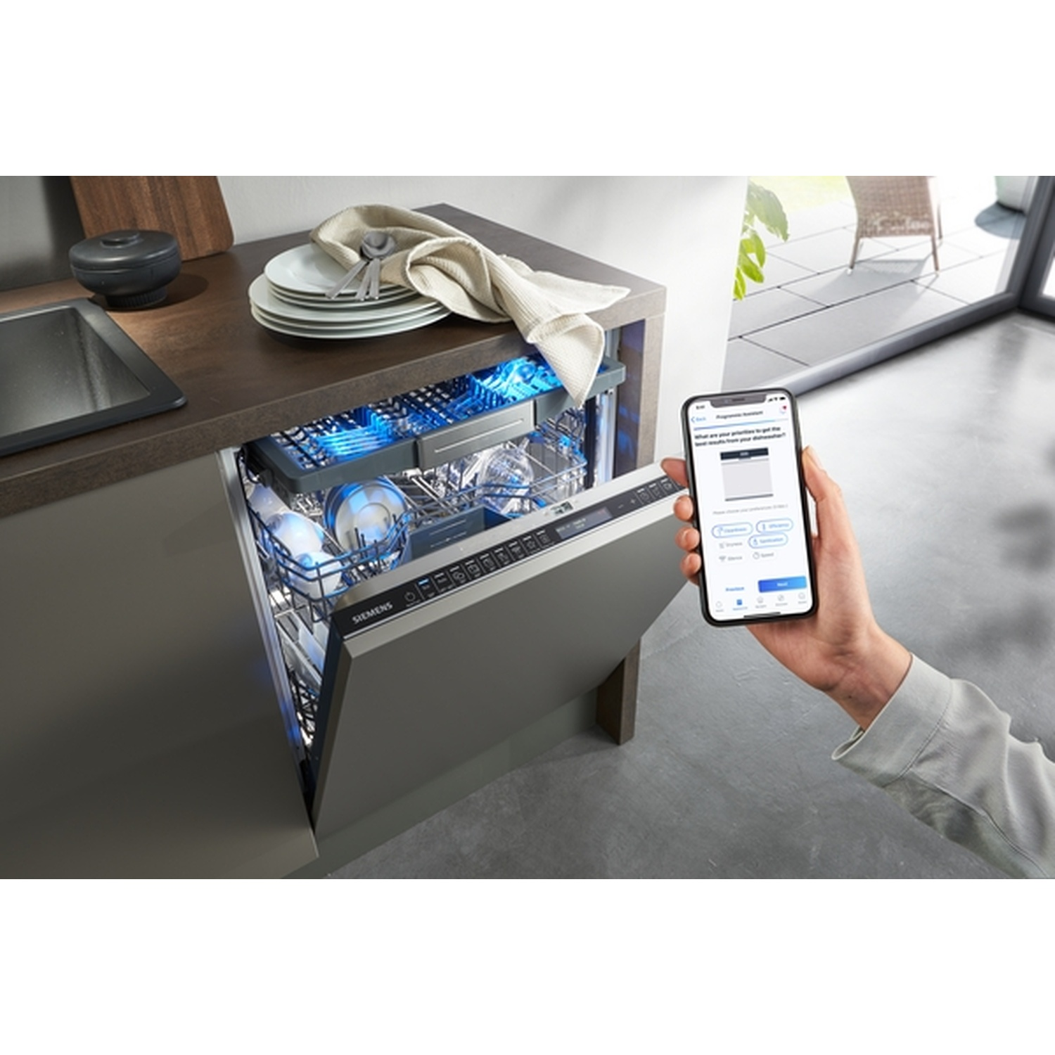 Lave-vaisselle avec porte ouverte, vaisselle à l'intérieur. Une main tient un smartphone avec une interface d'application. Assiettes et une serviette.