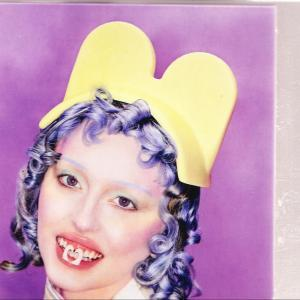 Eine Frau mit blauen Haaren und einem gelben Hut lächelt, mit einem dekorativen Objekt im Mund, vor lila Hintergrund.