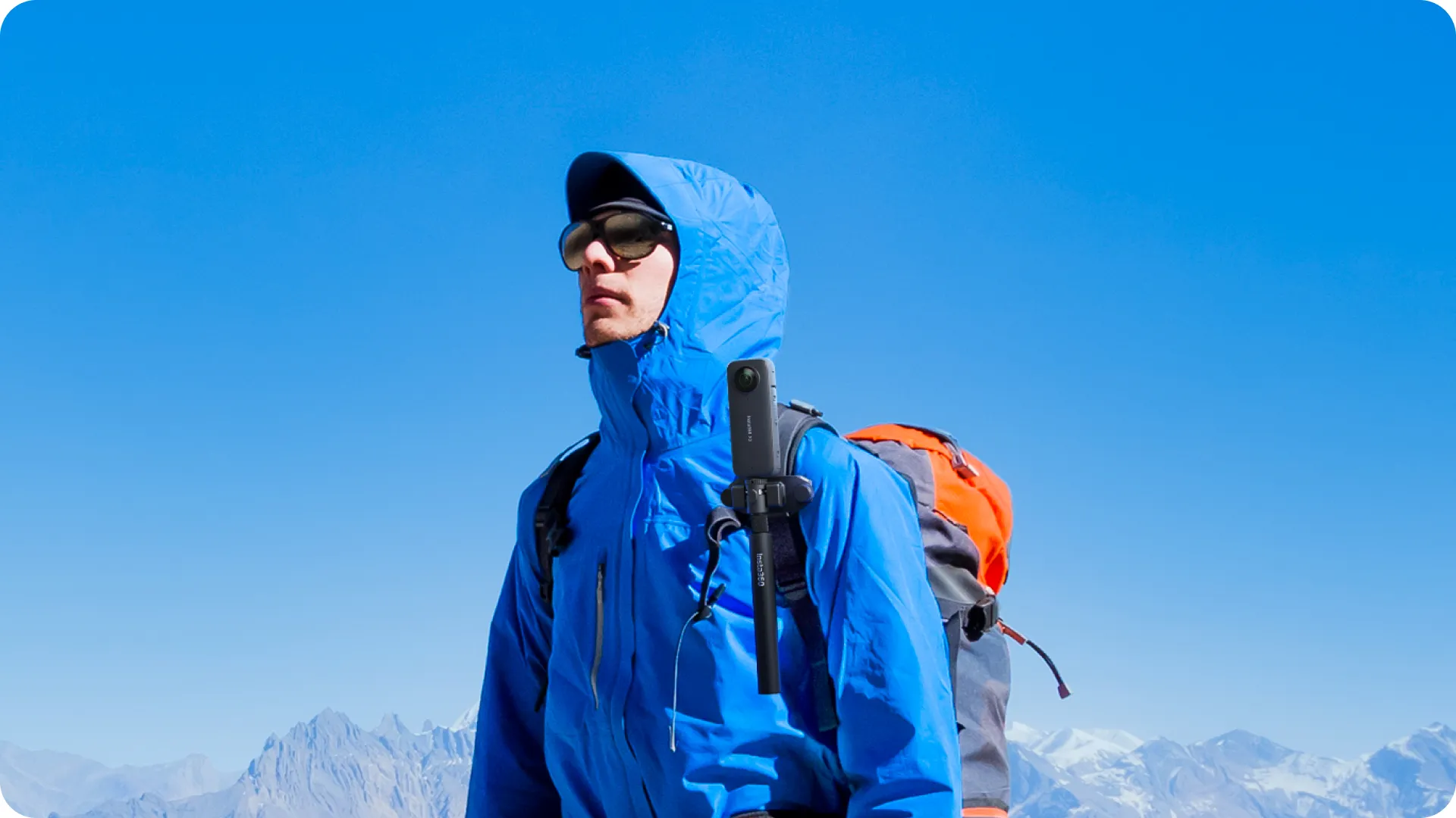 Mann in blauer Jacke mit Rucksack und Sonnenbrille, hält eine Kamera an einem Stock, vor Bergkette und blauem Himmel.