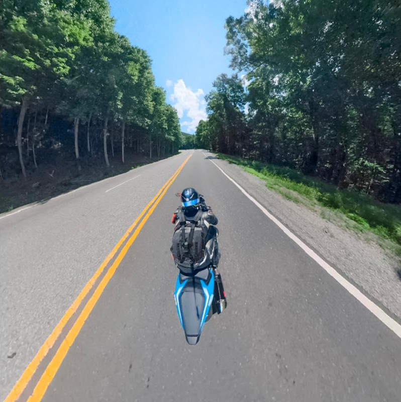 Un motocycliste roule sur une route droite, bordée d'arbres sous un ciel bleu. La route a des lignes jaunes.