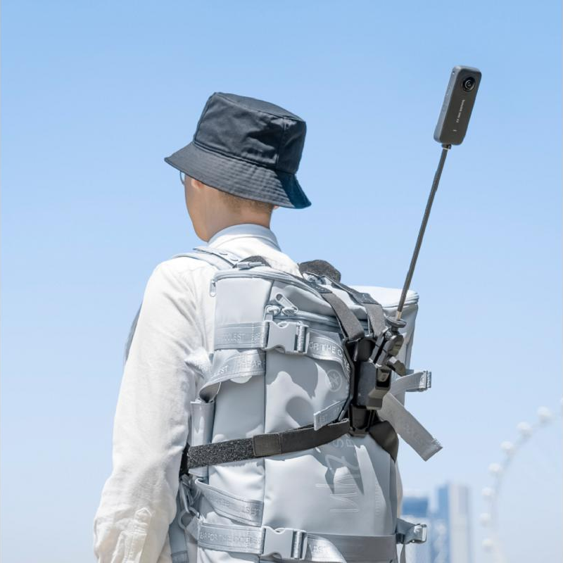 Une personne avec un sac à dos gris et une caméra. Elle porte un chapeau noir, dos à la caméra. Le ciel est bleu.