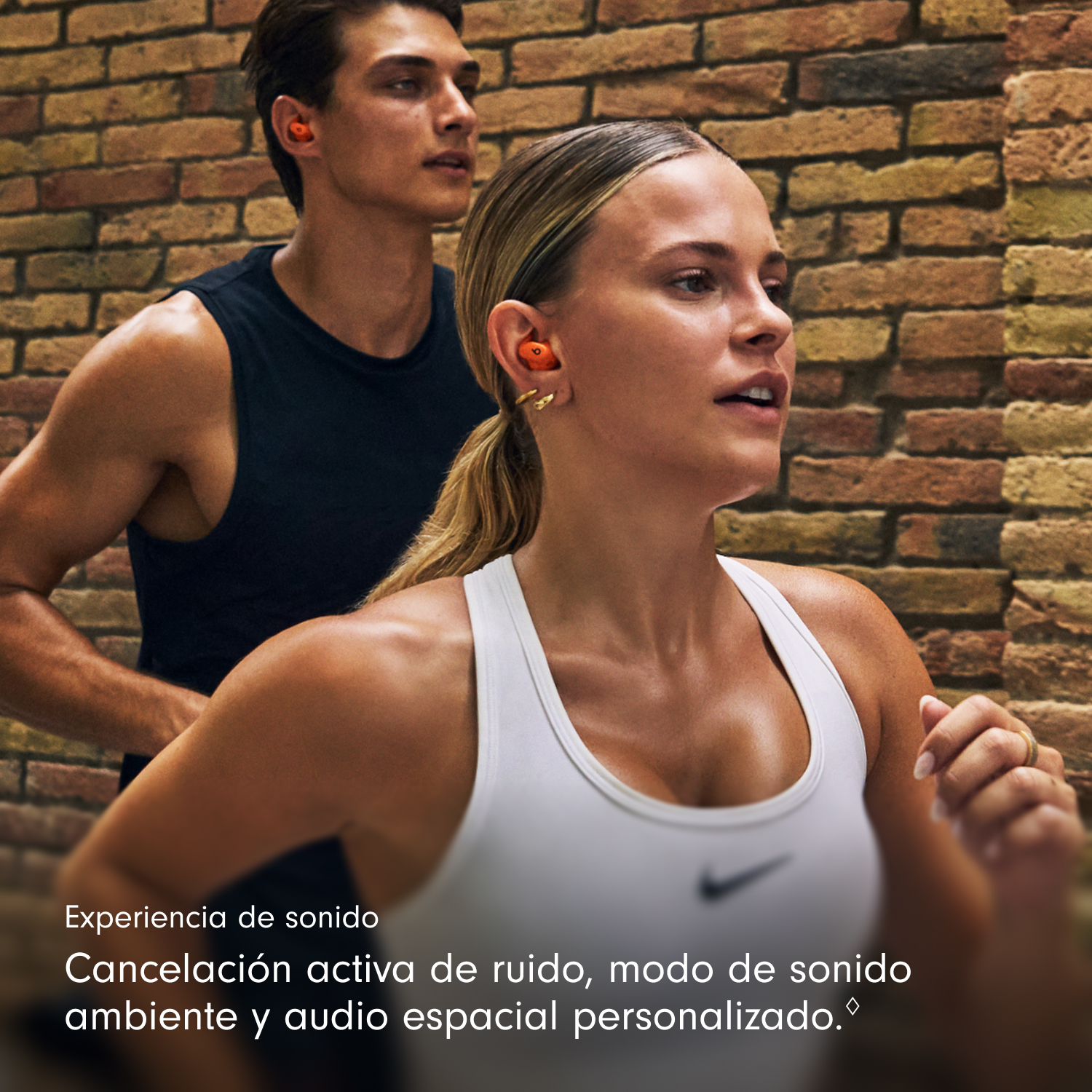 Un hombre y una mujer corren, ambos con auriculares naranjas. La mujer lleva una camiseta blanca. Un muro de ladrillos está al fondo.