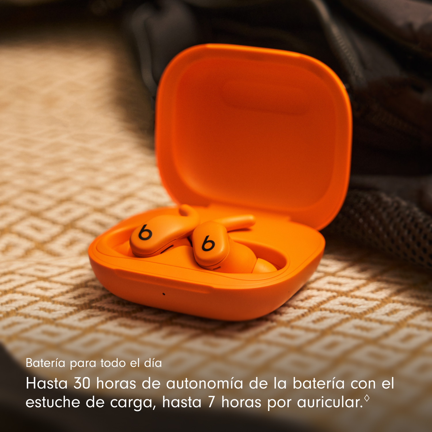 Estuche de auriculares naranja abierto con dos auriculares dentro. El fondo es una superficie estampada y una bolsa oscura.