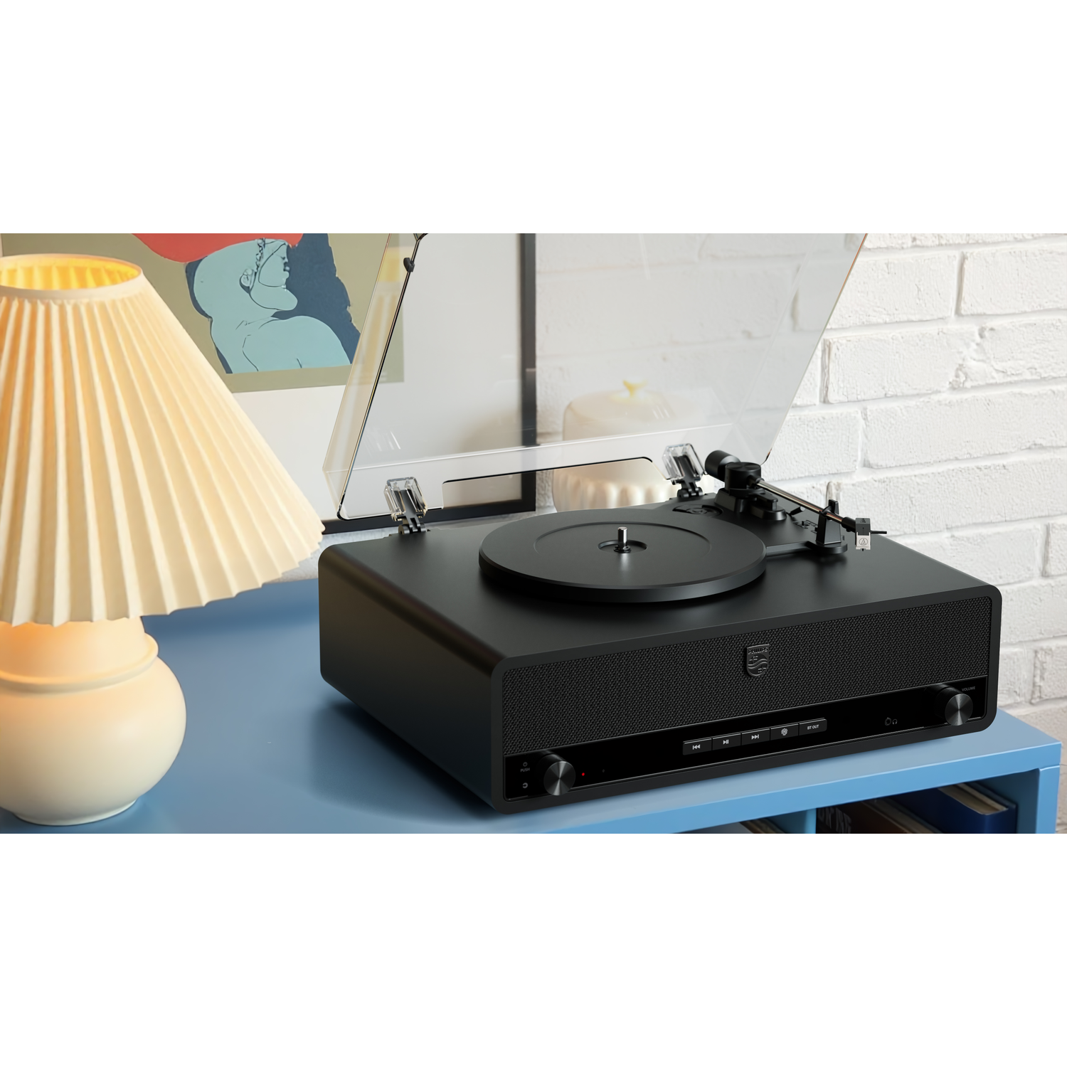 A black record player with a clear cover sits on a blue table. A lamp and a picture are beside it. A white brick wall is in the background.