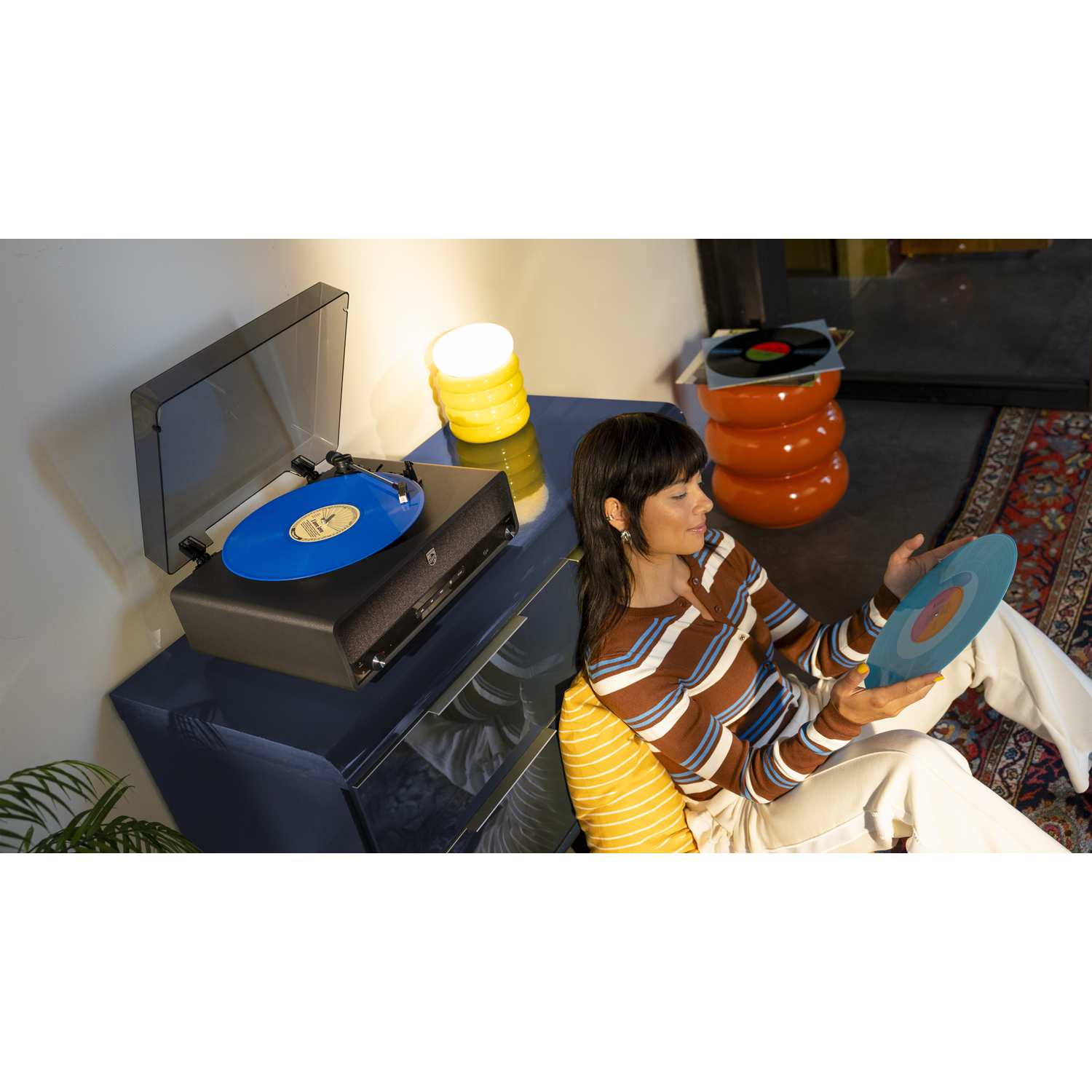 A woman holds a blue vinyl record, sitting near a turntable with another record. A yellow lamp and orange stool are visible.
