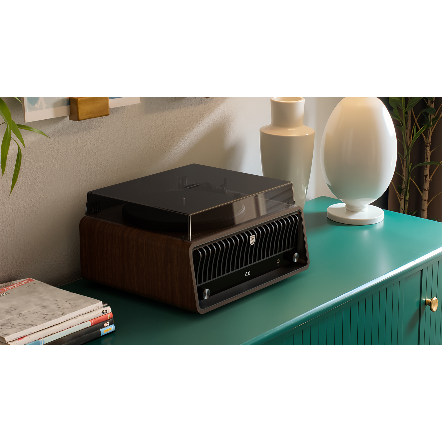 Een vintage platenspeler met een houten voet staat op een teal kast. Een witte vaas en lamp staan ernaast. Boeken zijn links opgestapeld.