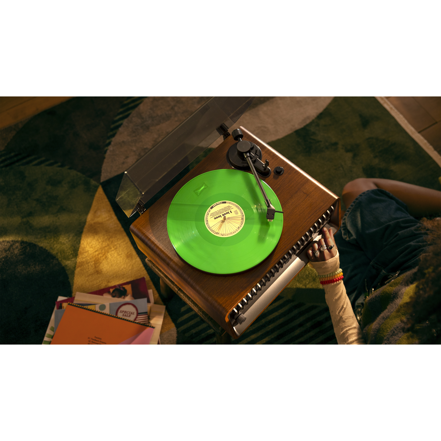 Een persoon ligt in de buurt van een platenspeler met een groene vinylplaat. Kleurrijke armbanden en ringen sieren hun hand. Boeken liggen in de buurt verspreid.