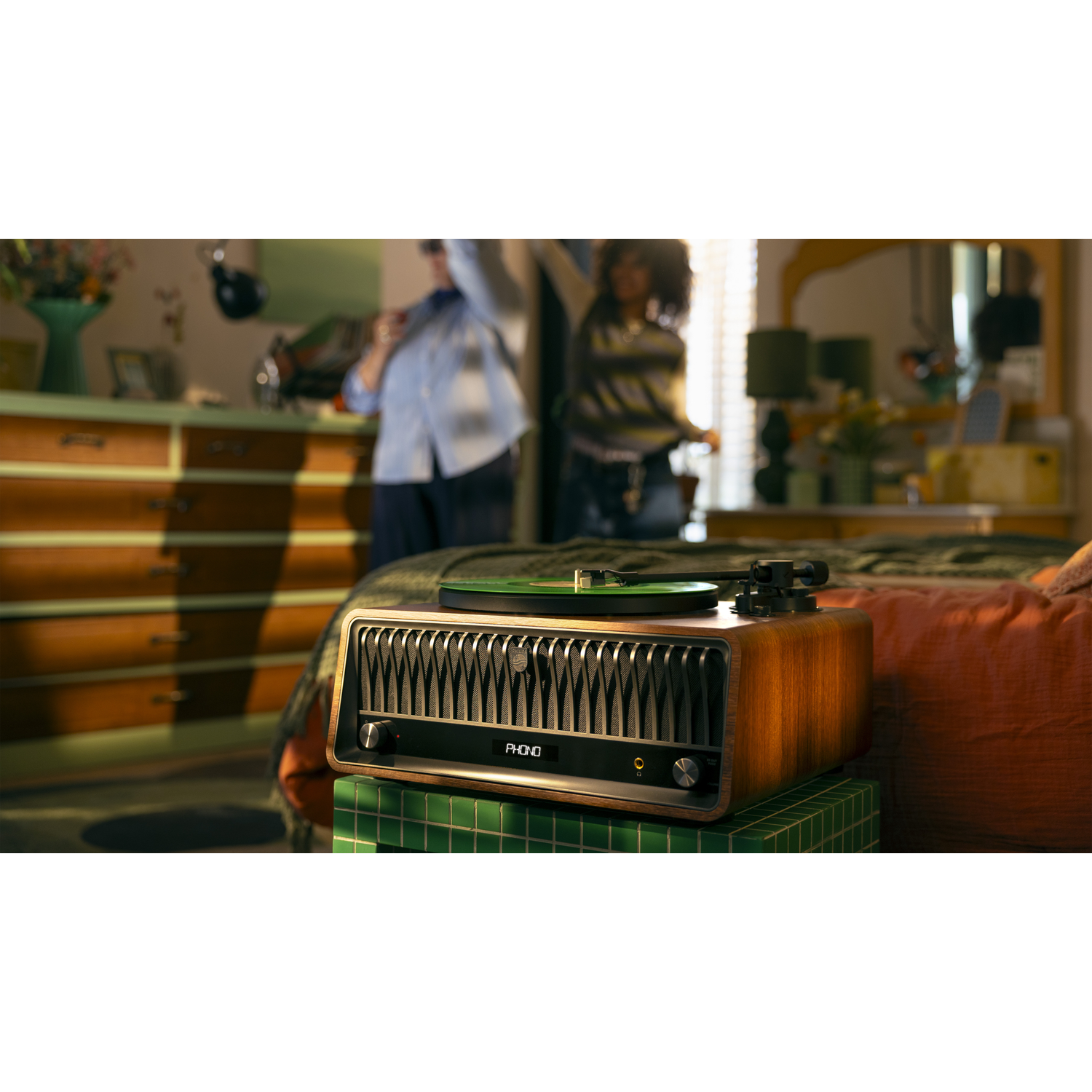 Een platenspeler in retrostijl staat op een groene tegelblok. Mensen dansen op de wazige achtergrond. Warme verlichting en een slaapkameromgeving.