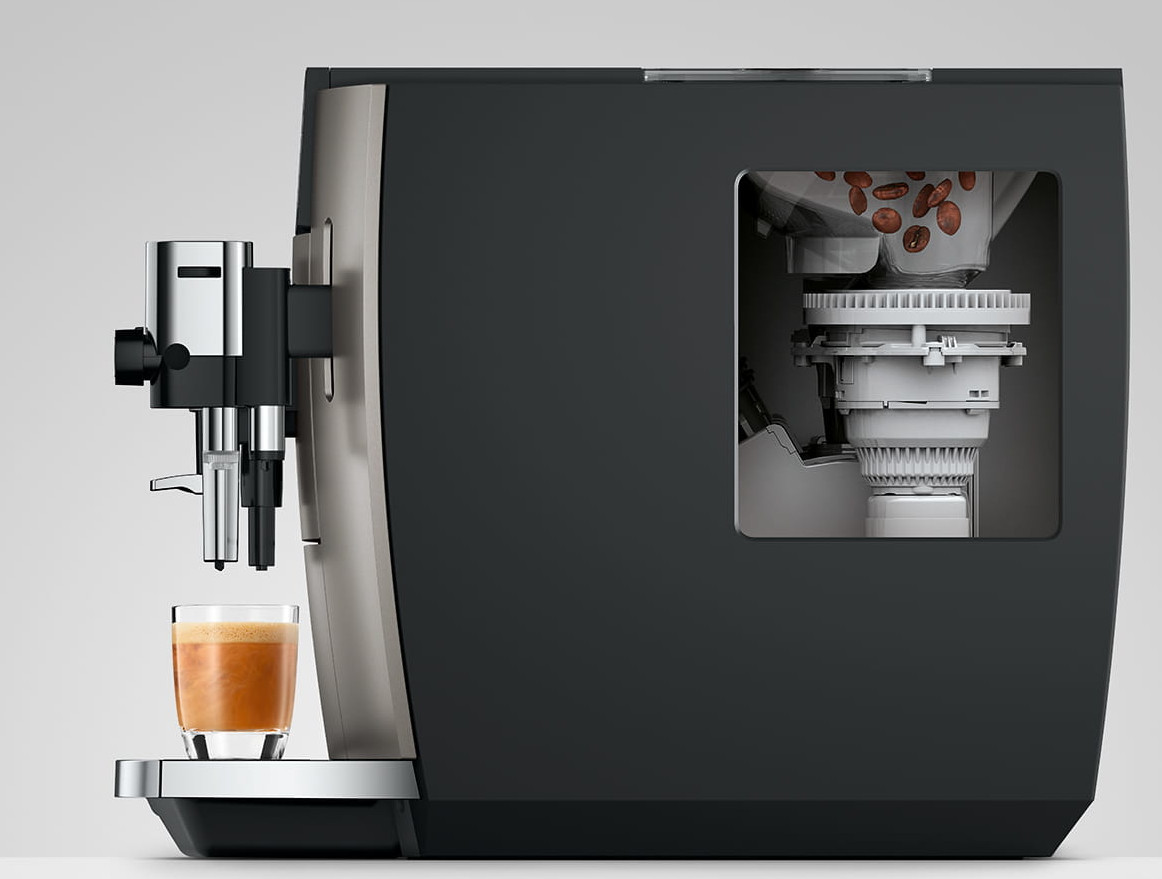 Ekspres do kawy ze szklanką kawy z przodu. W środku widoczne ziarna. Maszyna jest czarna i srebrna.