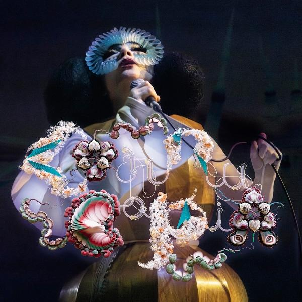 Bjork singt mit Mikrofon. Sie trägt eine blaue Maske und dekorative florale Elemente. Dunkler Hintergrund mit goldenem Kleid.