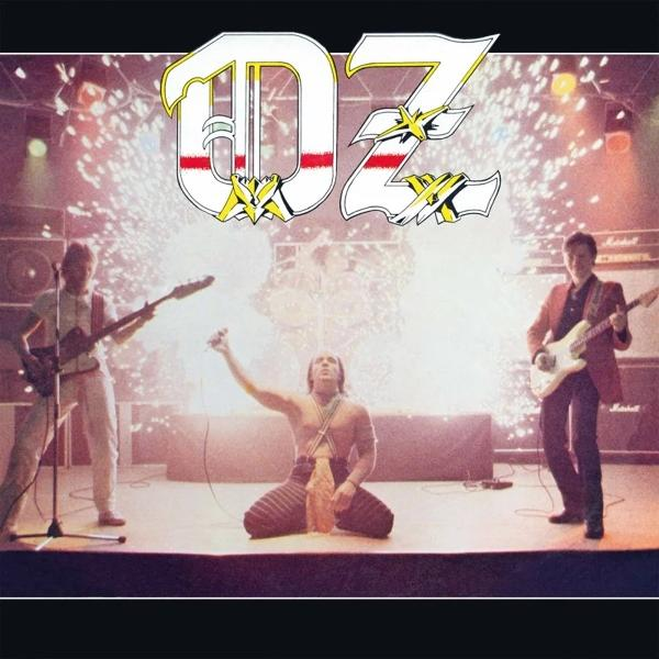 Band auf der Bühne. Ein Mann kniet, ohne Hemd. Zwei Gitarristen stehen. Der Bandname 'OZ' steht oben. Feuerwerk im Hintergrund.