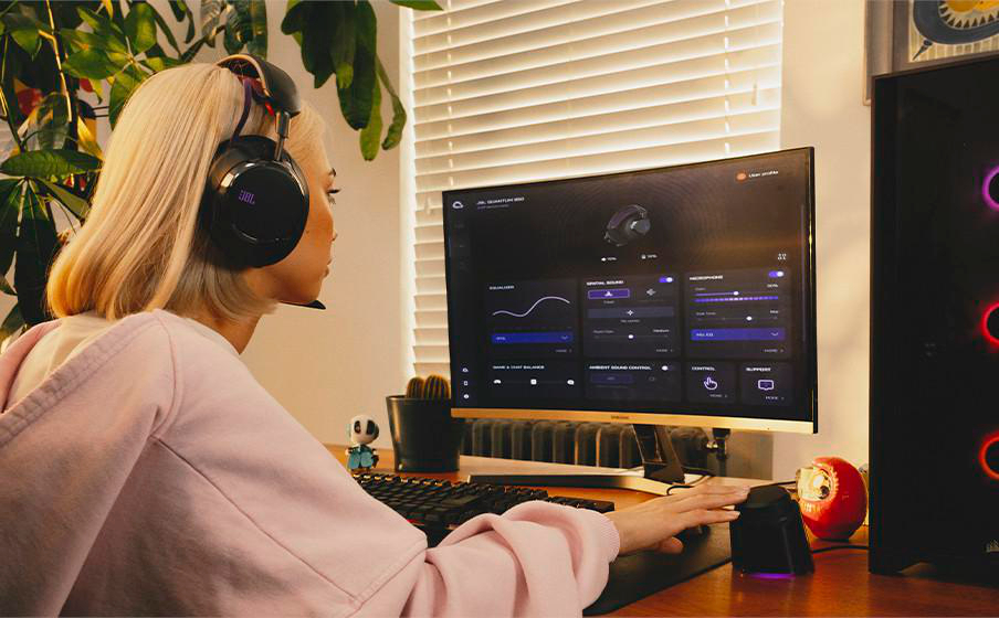 Eine Frau mit Kopfhörern sitzt an einem Computer. Sie passt die Einstellungen am Monitor an. Ein Gaming-PC und Zubehör sind vorhanden.