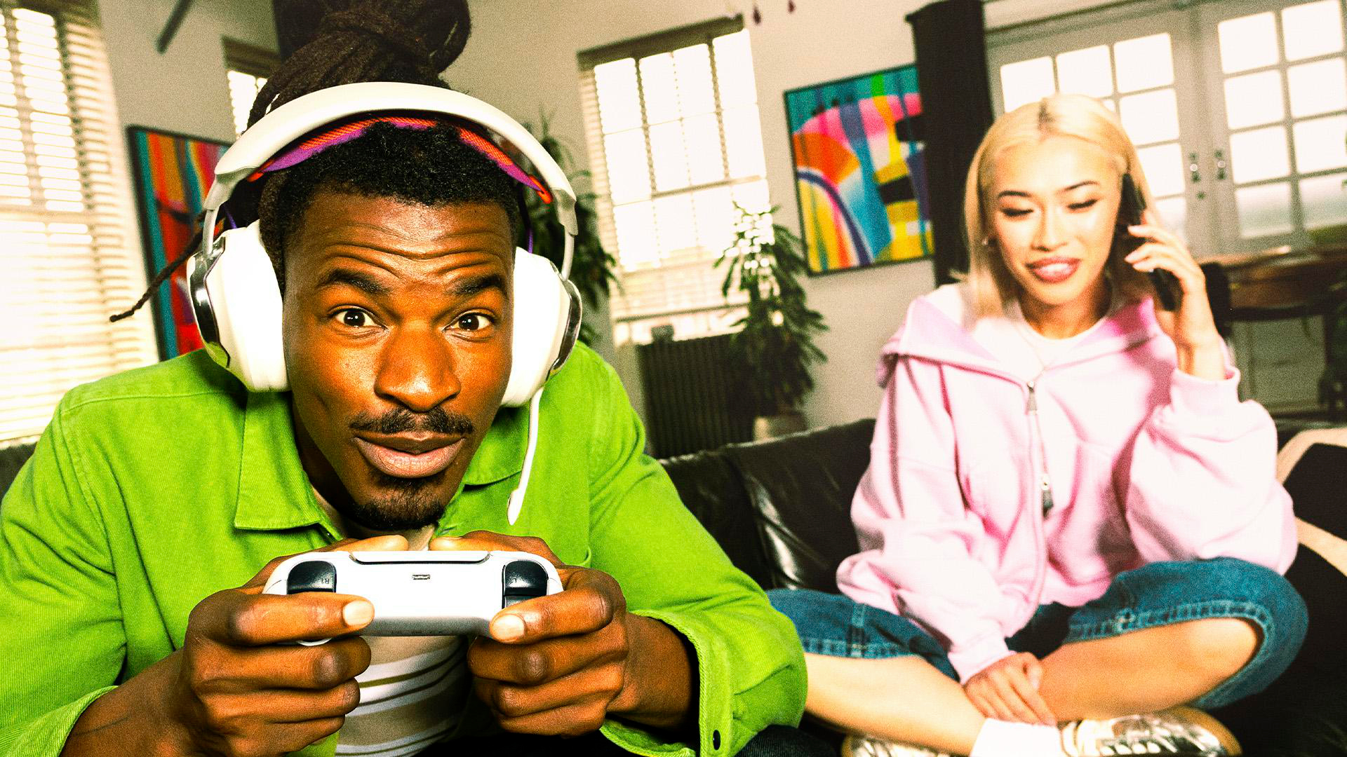 Ein Mann mit Kopfhörern spielt ein Spiel, während eine Frau sitzt und telefoniert. Sie sitzen auf einem schwarzen Sofa im Wohnzimmer.