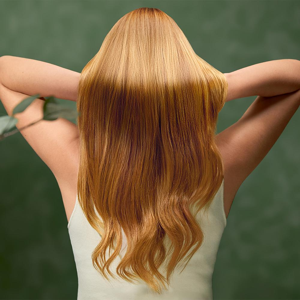 Mujer con cabello largo, rubio rojizo, brazos detrás de la cabeza. Con camiseta sin mangas blanca, fondo verde, hojas en la esquina izquierda.
