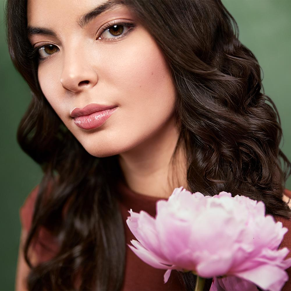 Una mujer de pelo oscuro y ojos marrones mira a la cámara. Una flor rosa está en primer plano. Fondo verde.
