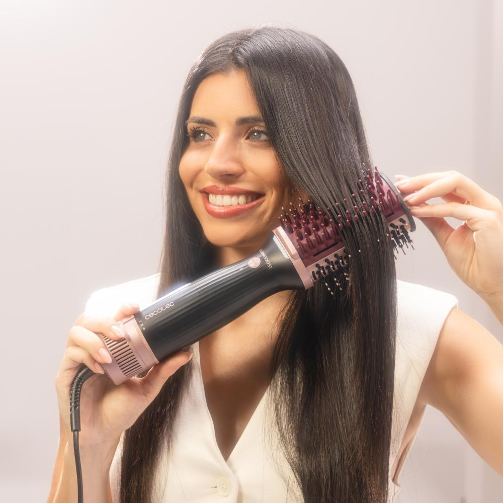 Una mujer sonríe mientras usa una herramienta negra y oro rosa para peinar su largo cabello oscuro. Lleva una blusa blanca.