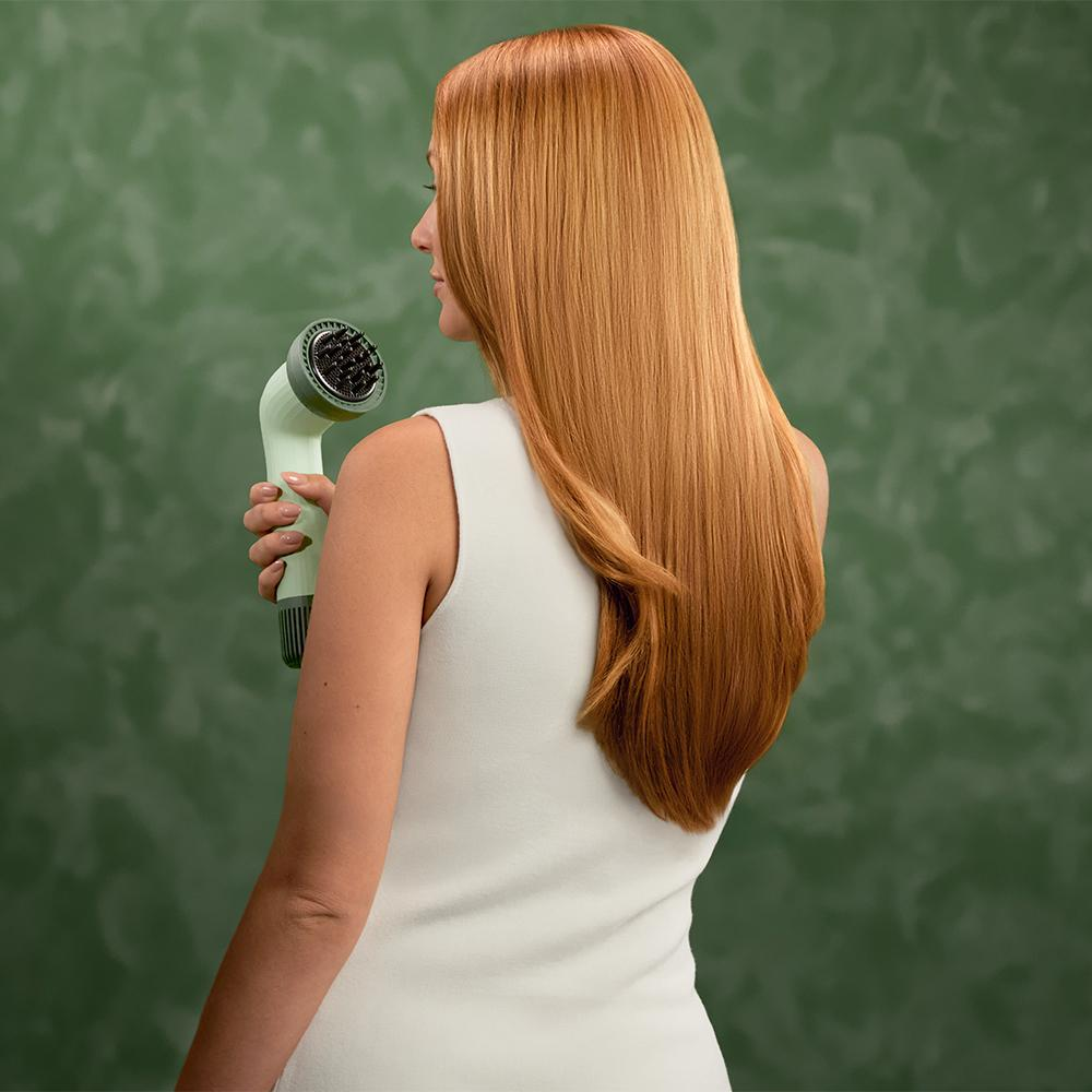 Mujer sosteniendo secador de pelo. Pelo largo rojo, top blanco, fondo verde.