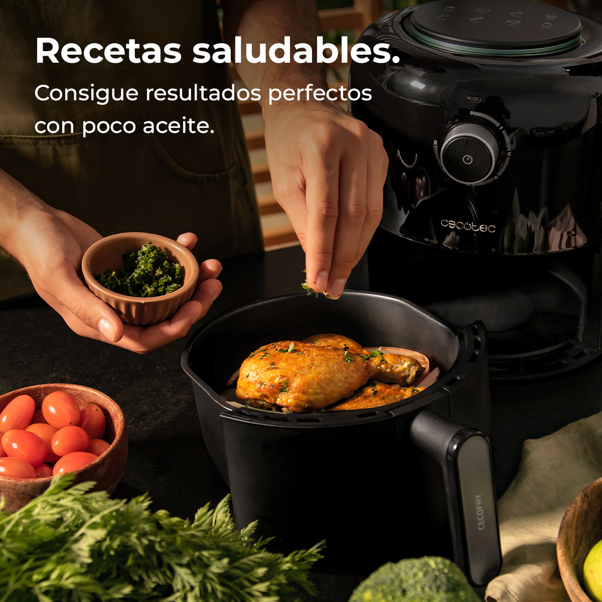 Una persona espolvorea hierbas sobre pollo en una freidora de aire. Tomates, hierbas y freidora de aire sobre encimera oscura.