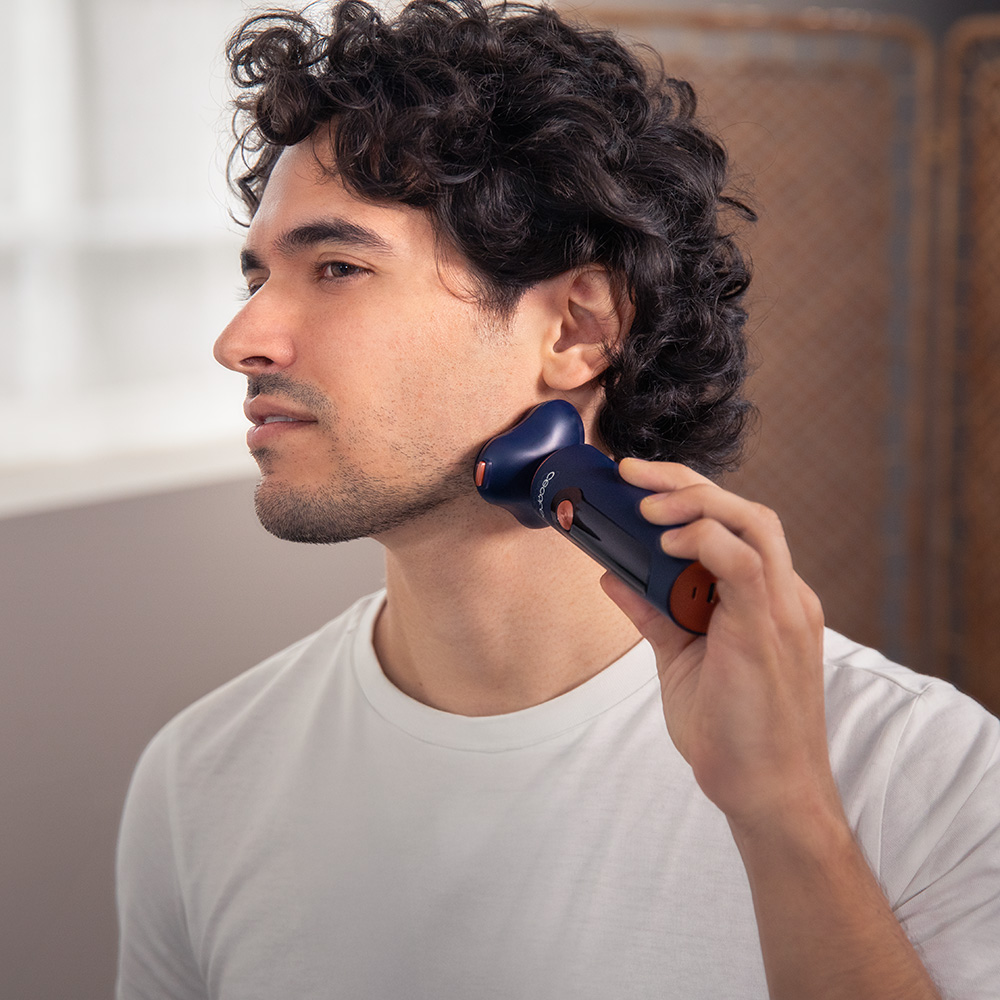 Un hombre se afeita con una afeitadora eléctrica azul y naranja. Usa una camiseta blanca. Se ve pelo rizado y barba incipiente.