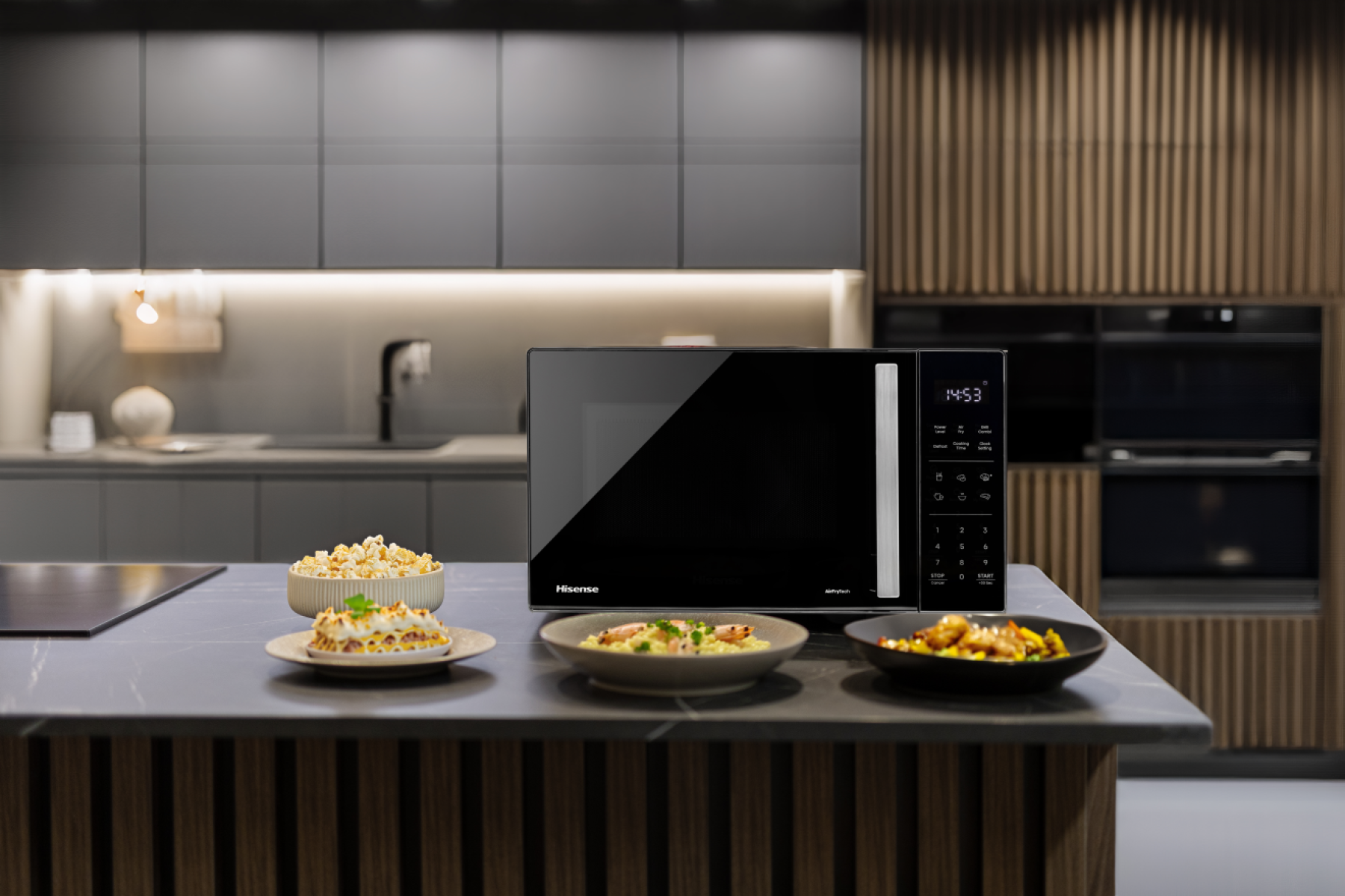 Eine schwarze Mikrowelle mit Essen davor in einer modernen Küche. Popcorn, Lasagne und Garnelen stehen auf der Theke.