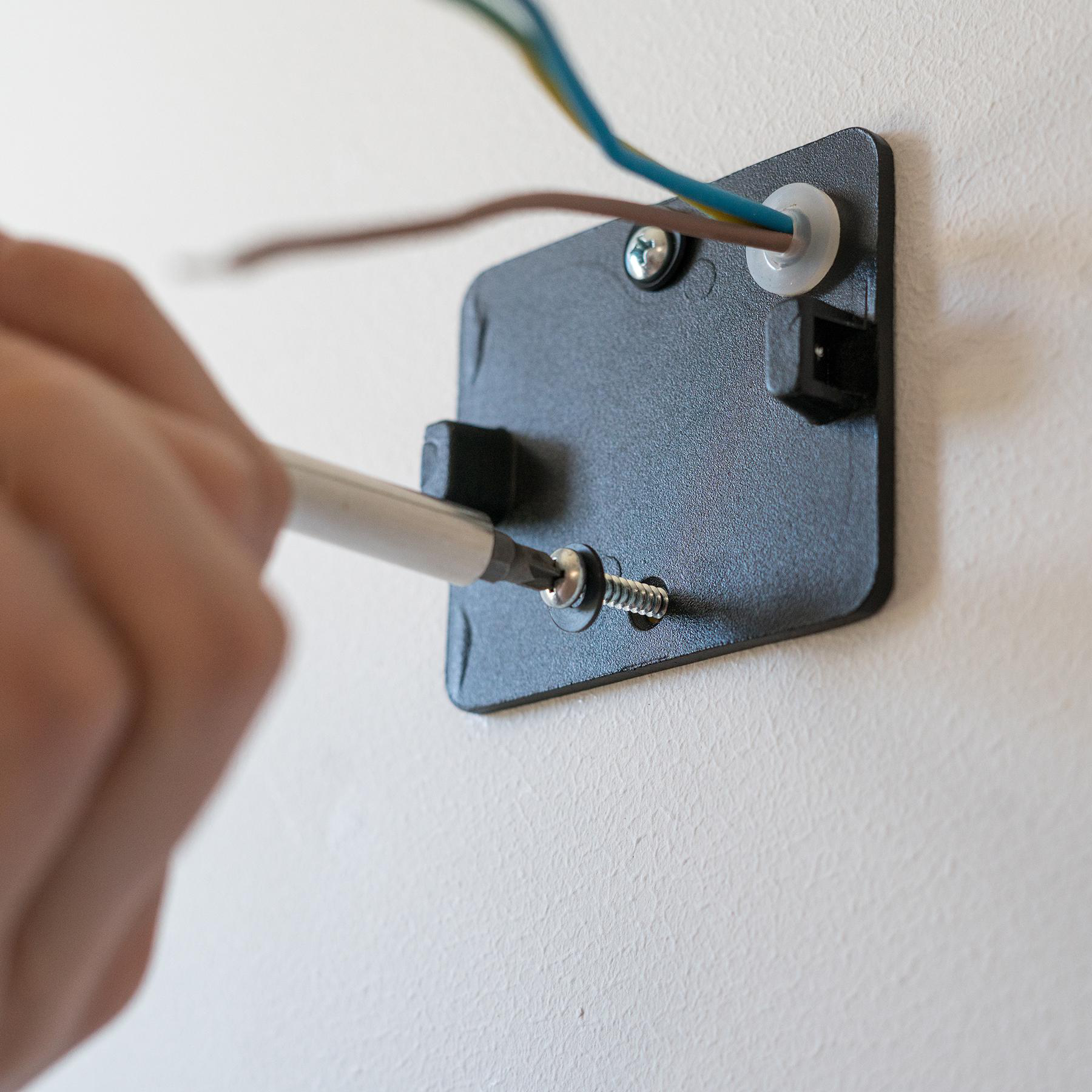Eine Hand, die mit einem Schraubendreher eine Schraube in eine schwarze Montageplatte mit Kabeln anzieht, vor einer weißen Wand.