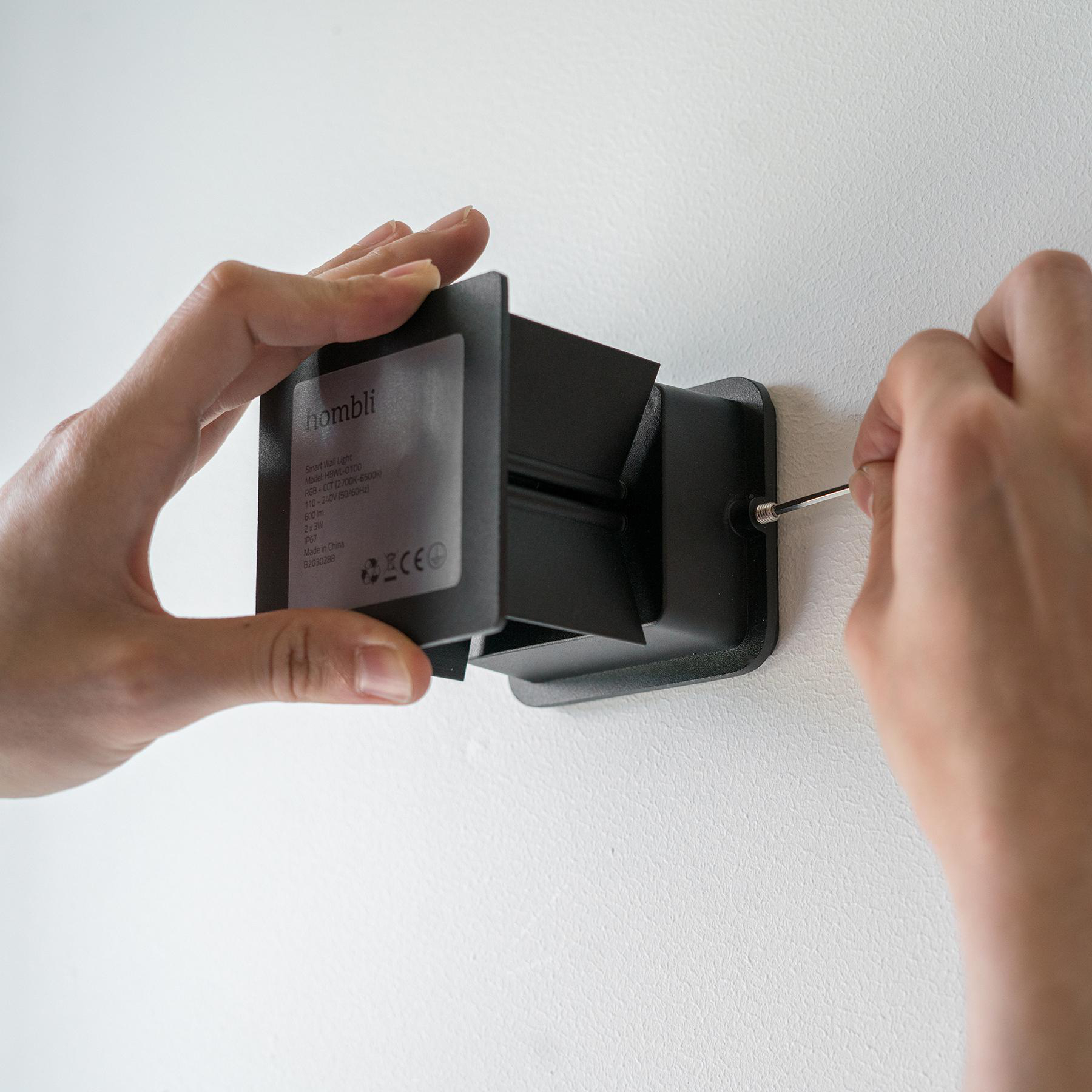 Hände montieren eine schwarze Wandleuchte an einer weißen Wand. Ein Schraubendreher wird benutzt.