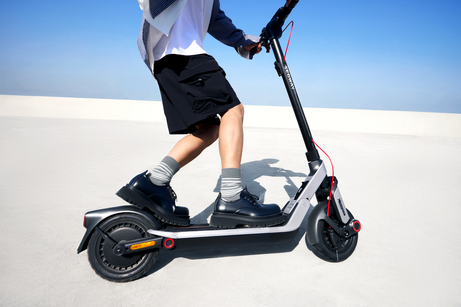 Una persona monta un patinete Segway. Lleva pantalones cortos negros, zapatos y calcetines a rayas. El fondo es blanco con cielo azul claro.
