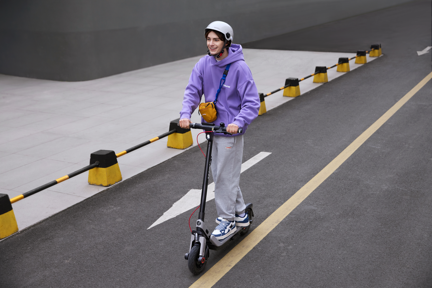 Una persona con una sudadera con capucha morada monta un patinete eléctrico en una calle, usando un casco. El fondo incluye barreras viales y un edificio.