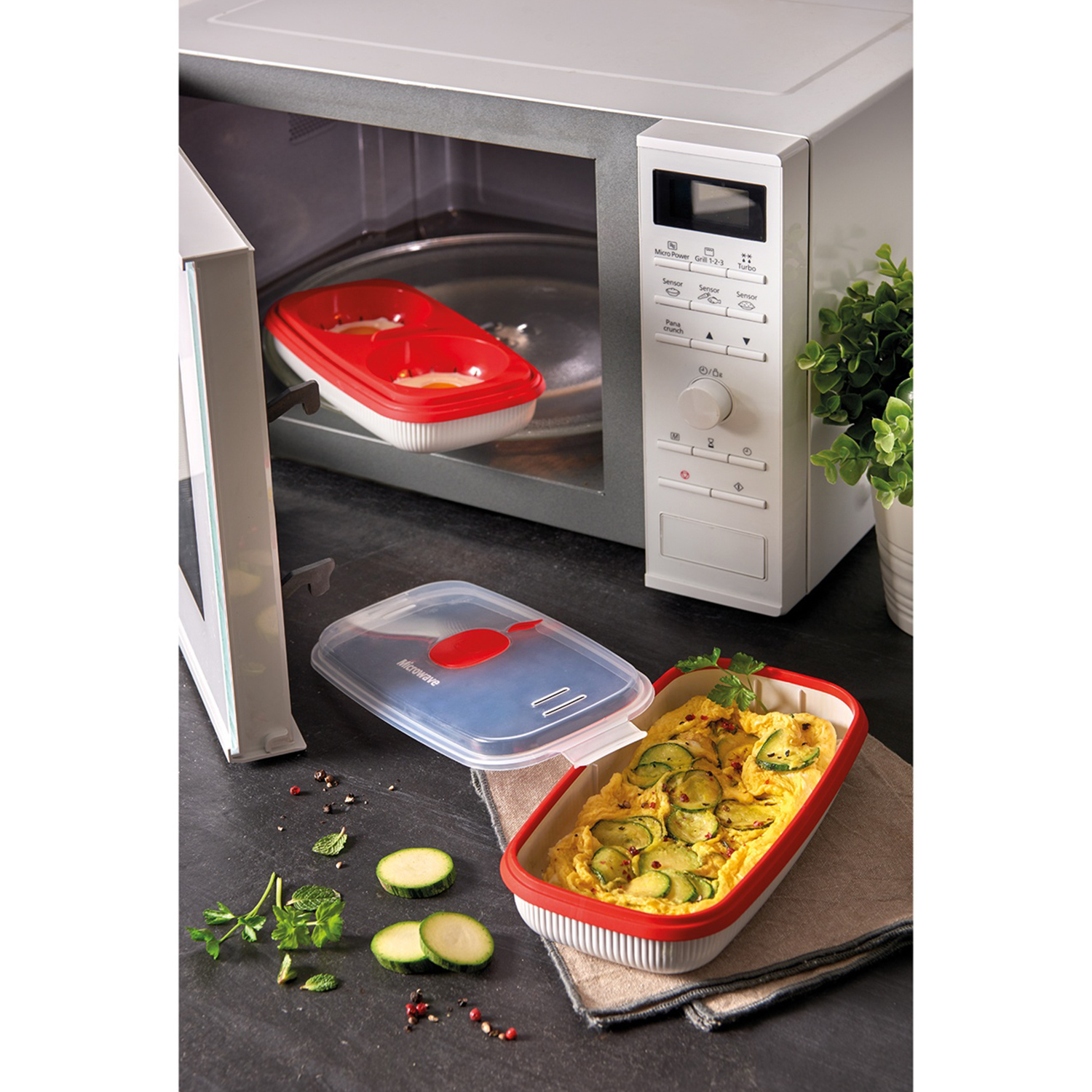 Microonde con porta aperta. All'interno, un contenitore rosso e bianco. Sul bancone, un contenitore con cibo, fette di zucchine ed erbe aromatiche.