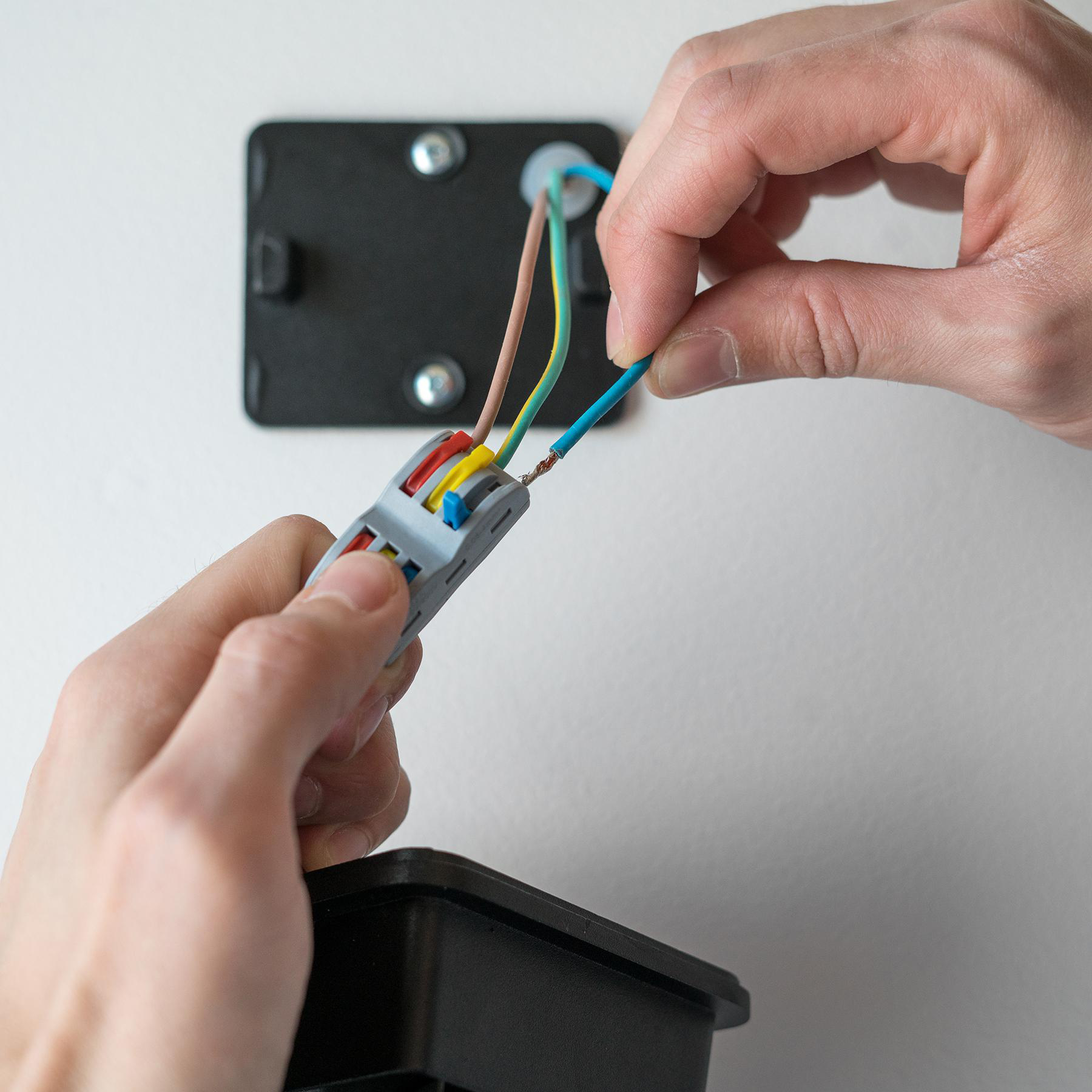 Hände verbinden Drähte mit einem Stecker. Drähte sind blau, braun, gelb und grün. Eine schwarze Box und Wand sind ebenfalls sichtbar.