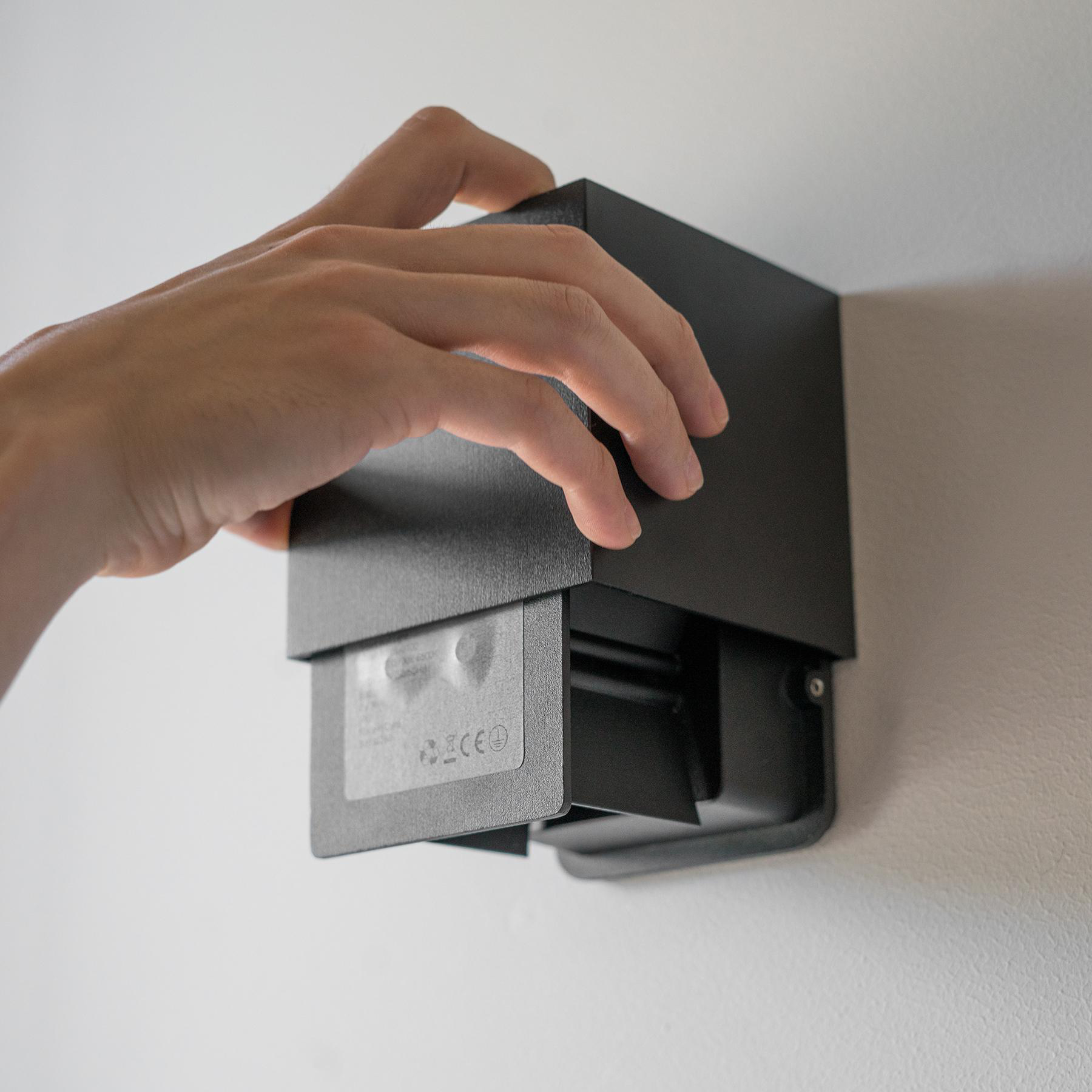 Eine Hand justiert eine schwarze, an der Wand befestigte Leuchte. Die Leuchte hat eine rechteckige Form und befindet sich an einer weißen Wand.