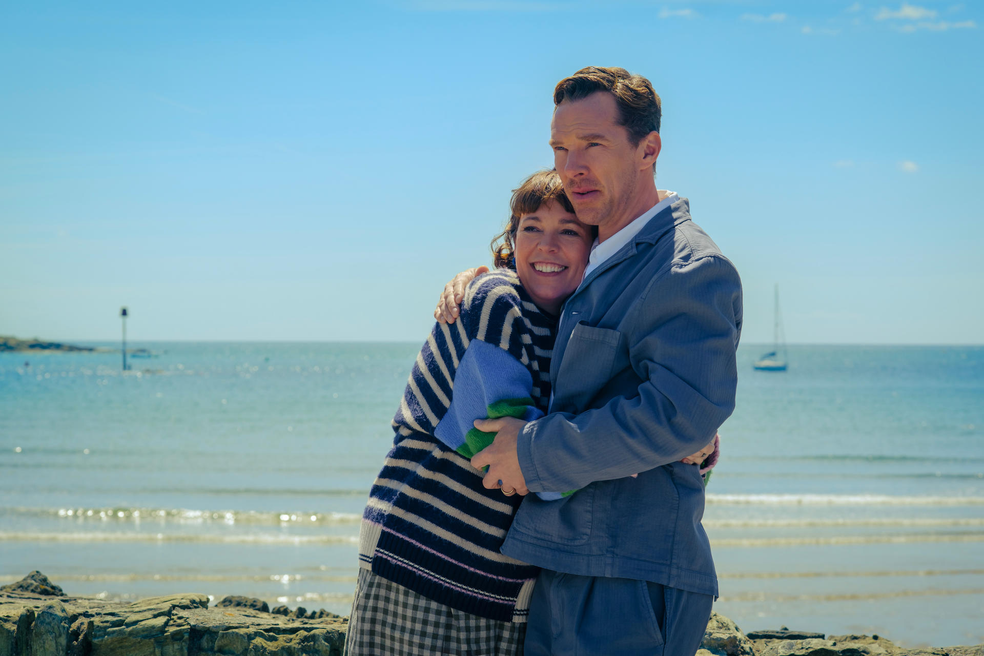 Zwei Personen umarmen sich am Strand. Frau in gestreiftem Pullover und Mann in grauem Anzug. Meer und Himmel im Hintergrund.
