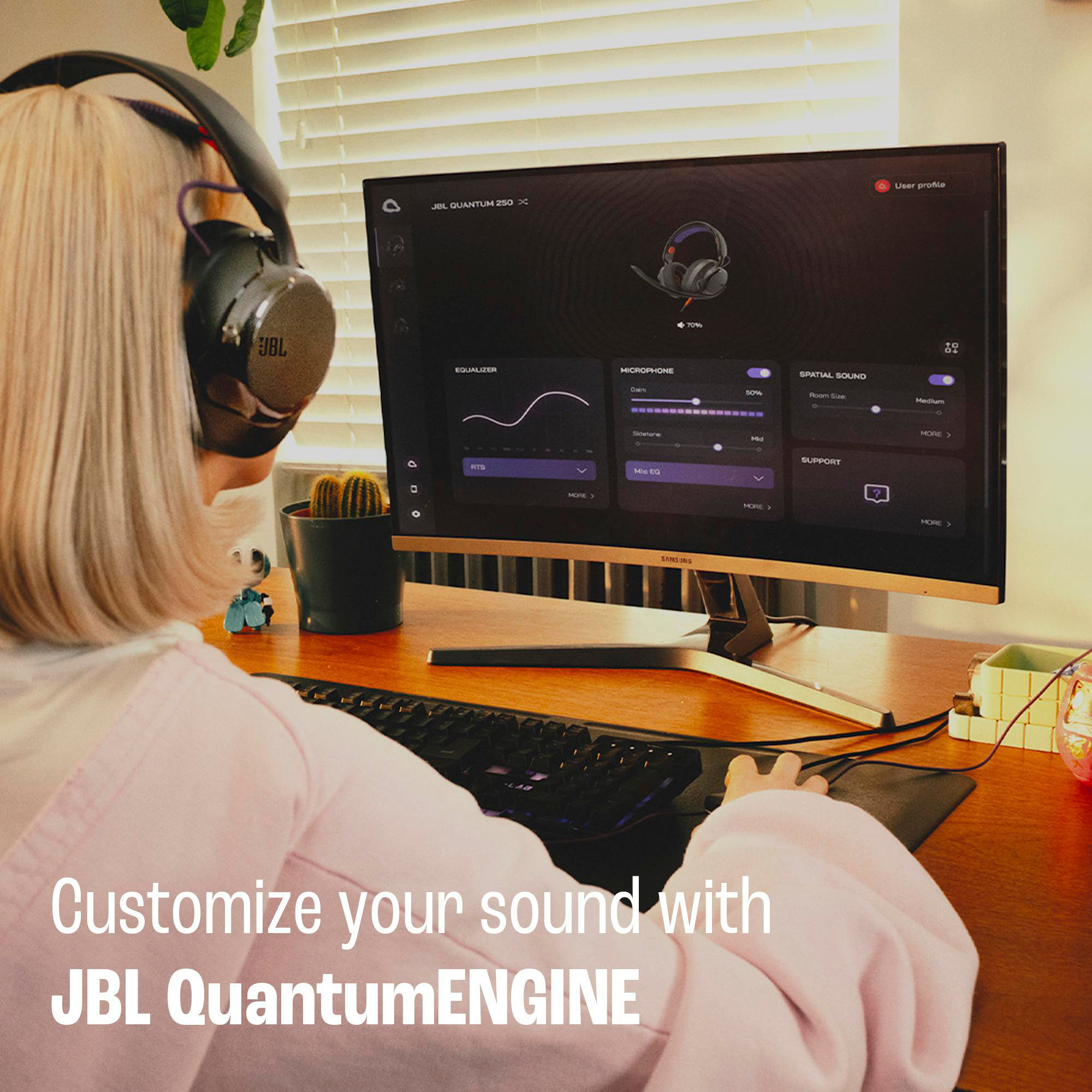 Eine Frau mit Kopfhörern passt Einstellungen auf einem Monitor an, der die JBL QuantumENGINE-Software anzeigt, Schreibtisch mit Tastatur und Kaktus.